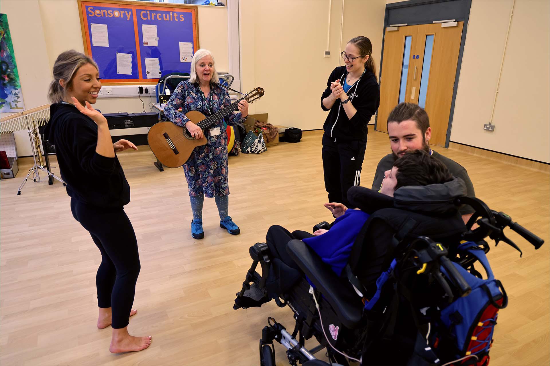 A person in a wheelchair looks on and enjoys the music played live on guitar while other people in the room look on and clap in time with the music