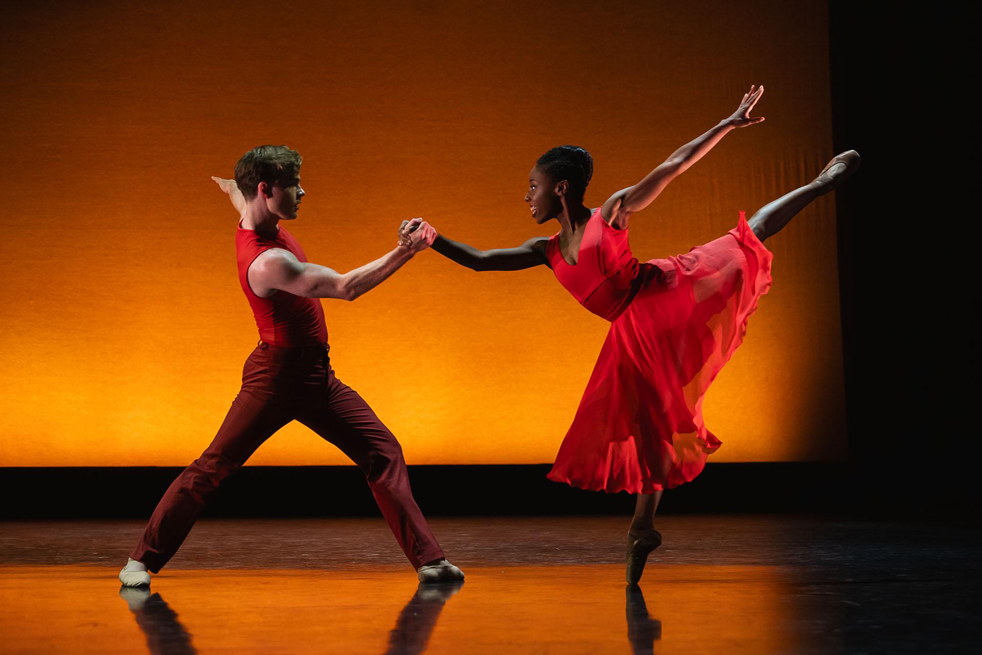 Woman balances en pointe on one foot, leg behind her raised high and another dancer helping her balance by holding her right hand