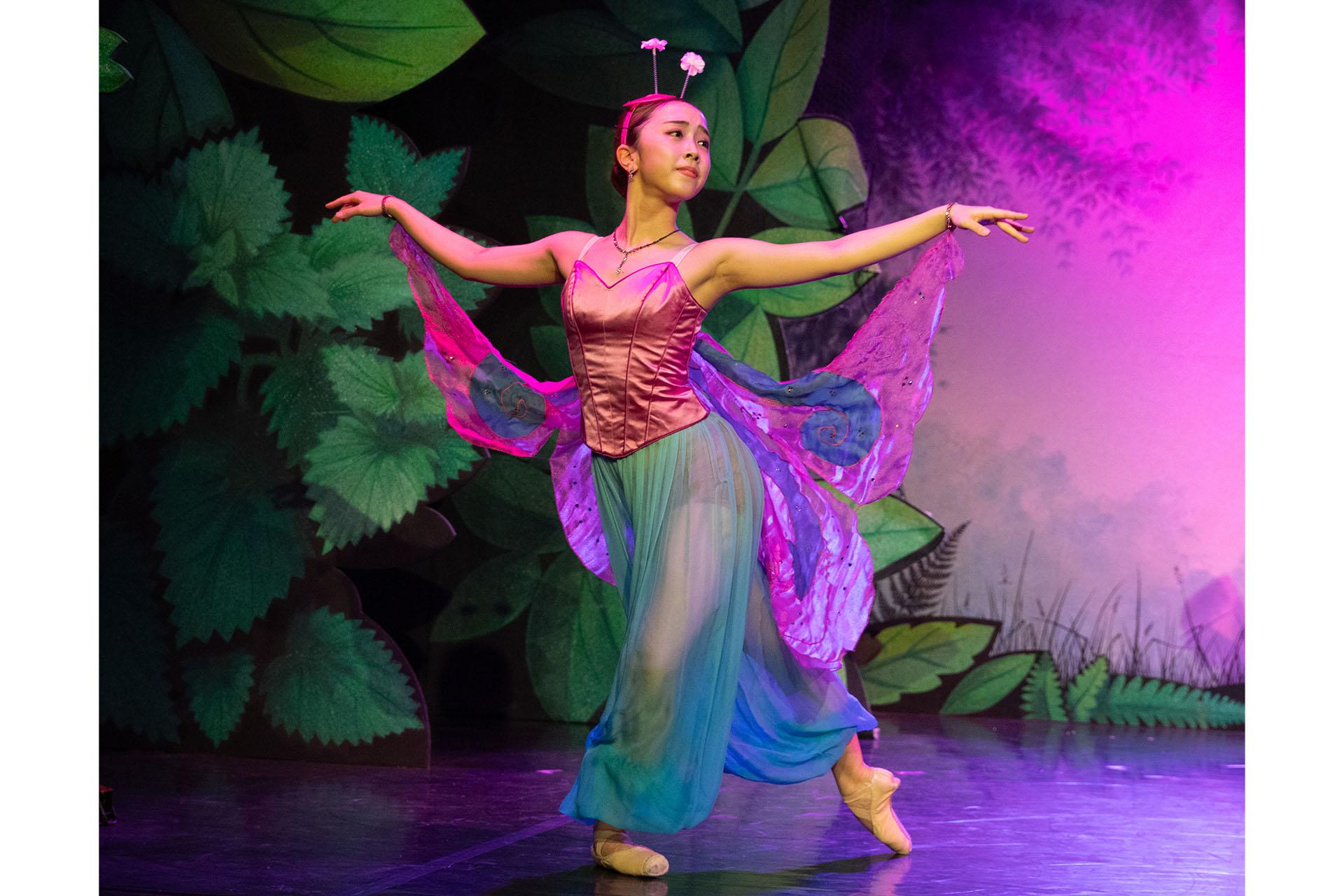 Female dancer, dressed as a butterfly in magenta and cyan, stands with her arms apart and looks stage left