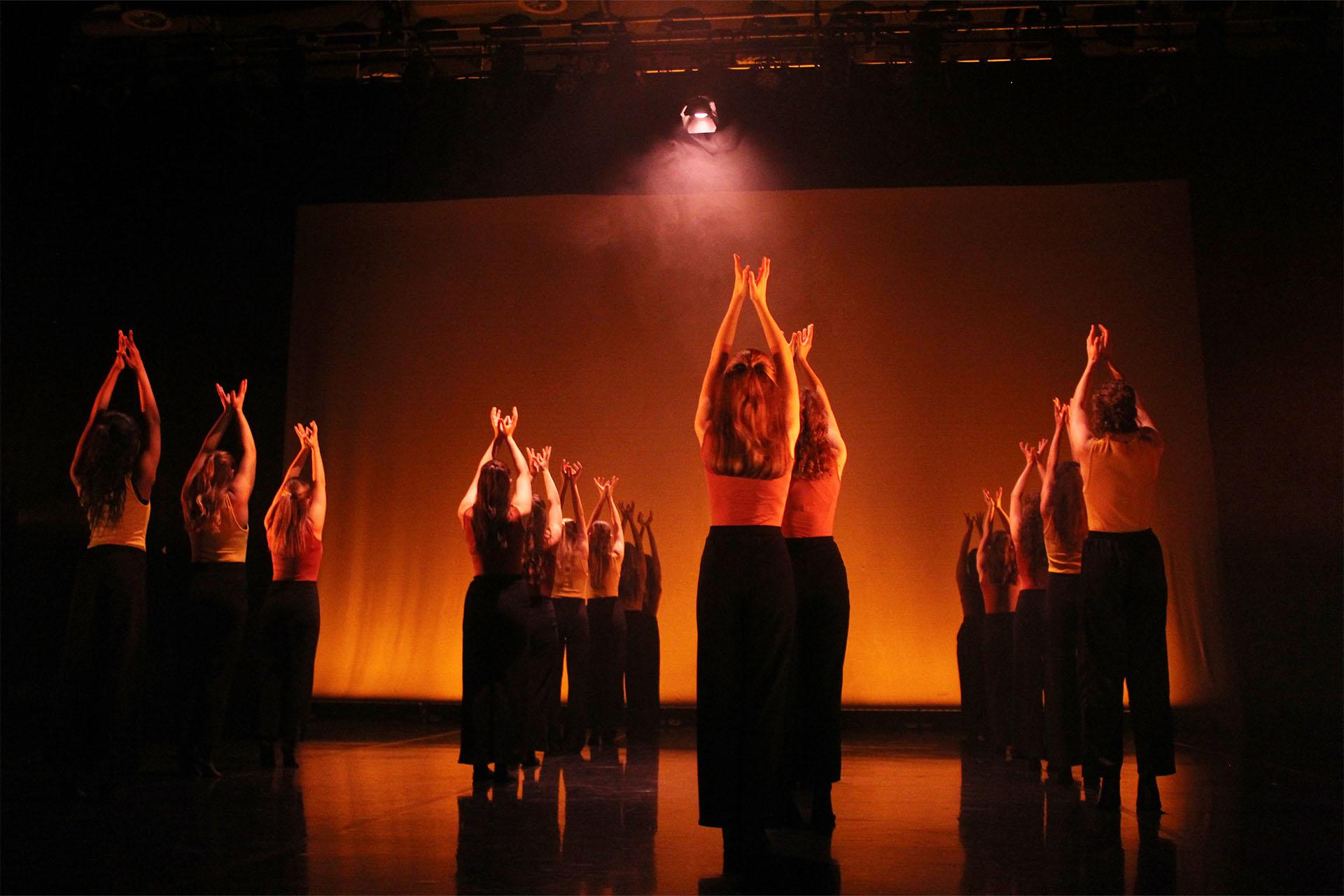 SABT, Yorkshire Youth Dance Platform, photo Edie Ashby Under orange light, a group of young dancers standing tall and reaching above their head.