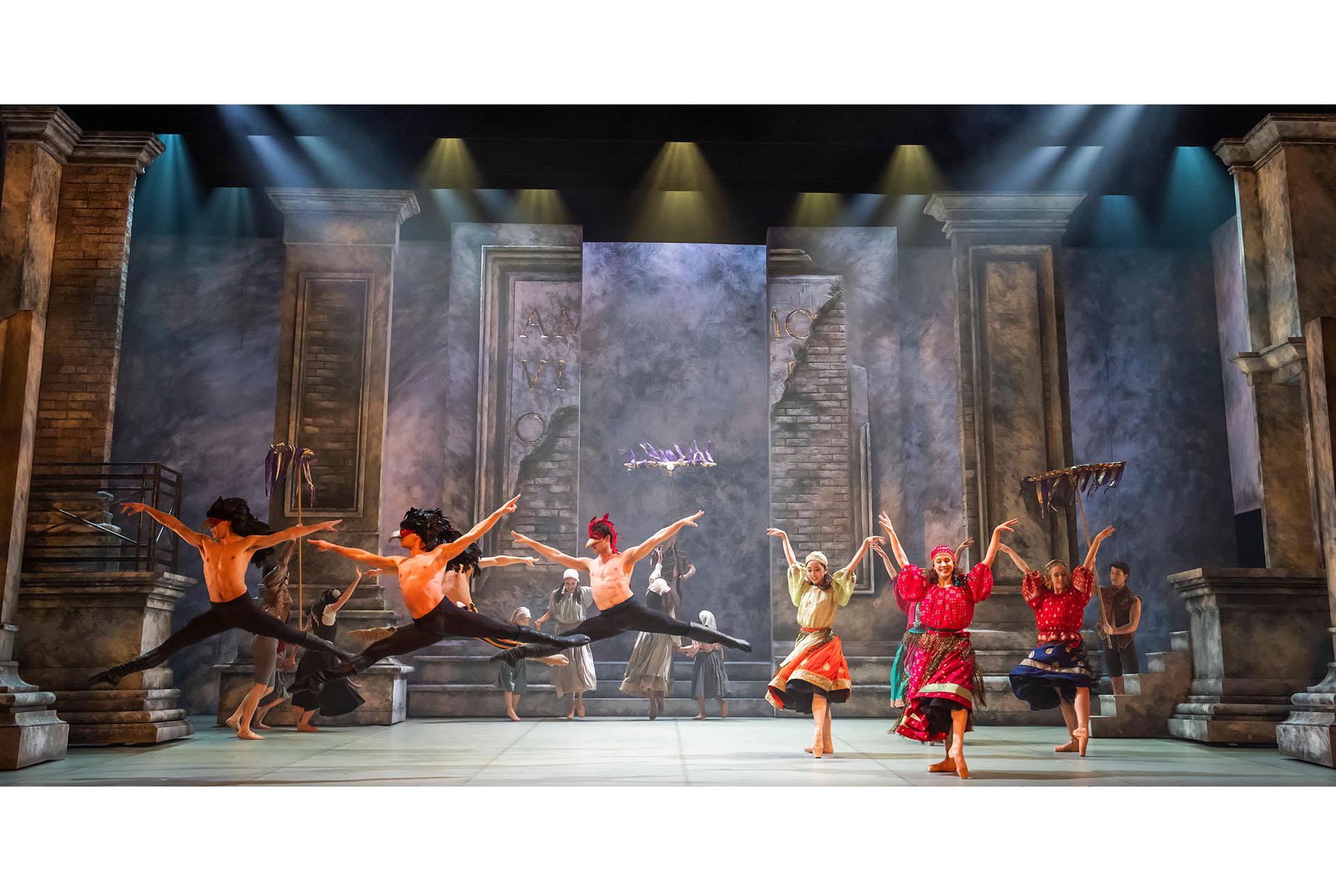 Northern Ballet dancers in Romeo and Juliet. Photo Tristram Kenton Three dancers on the left of a large stage in bird masks. To the right is a group of ladies in bright skirts.
