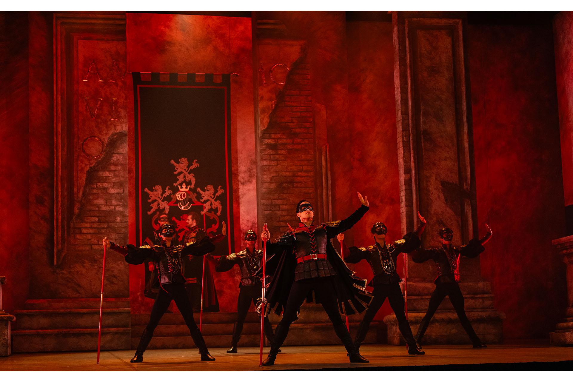 I red lighting, a group of men stand wearing masks and carrying a red staff