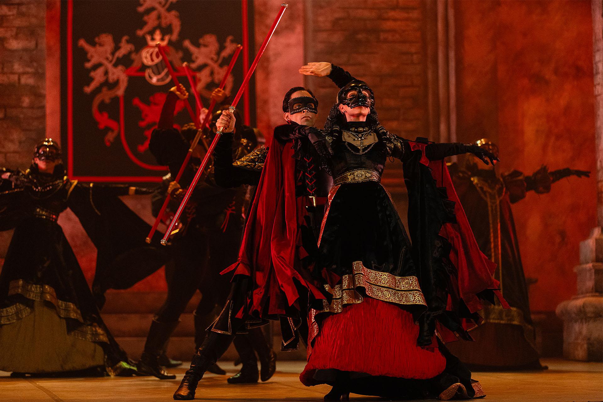 Dressed in red and black and wearing masks, Lord and Lady Capulet stand imperiously facing out from the stage