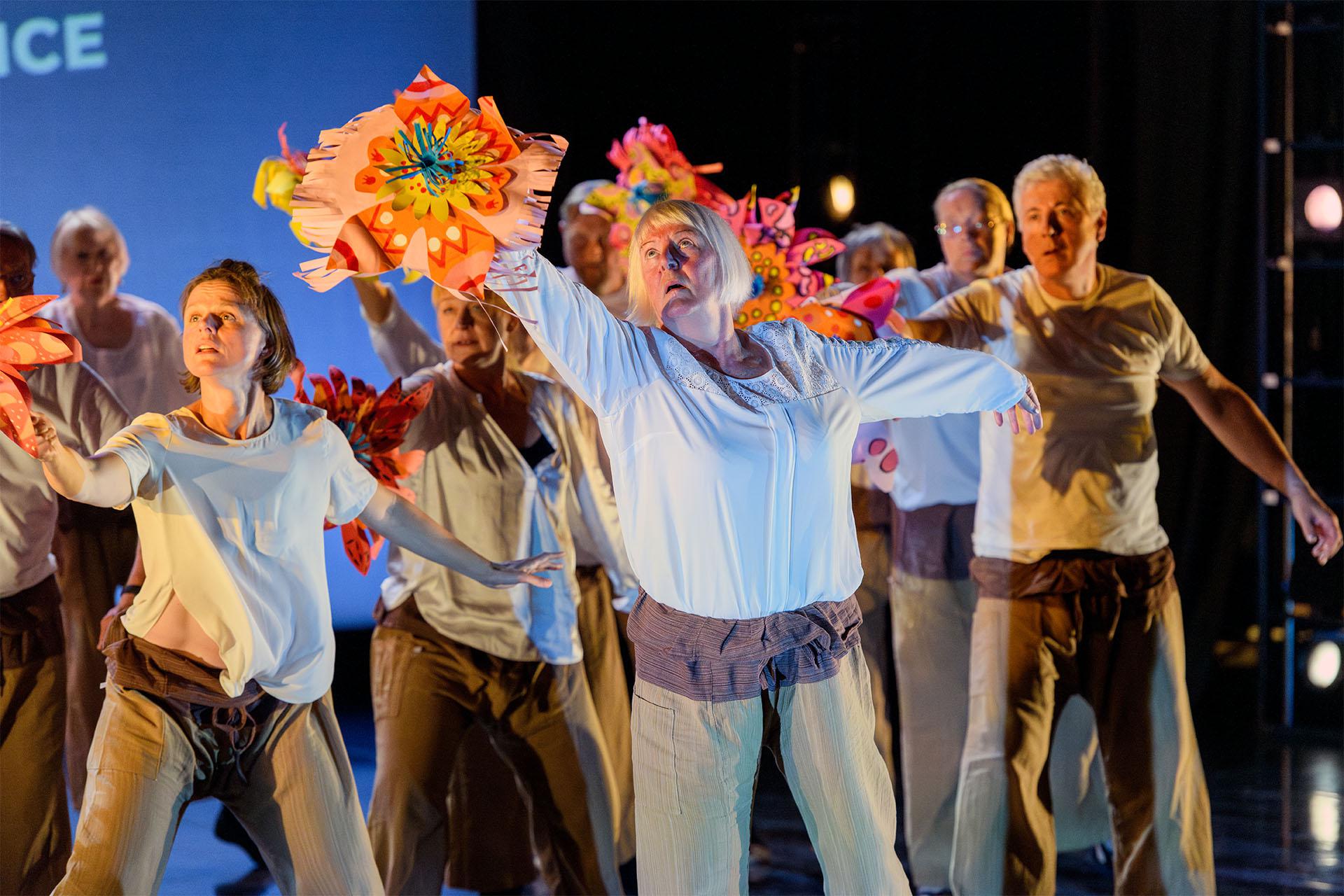 Group of people wearing similar clothes look to their right, arms spread, one hand each holding a bright flower