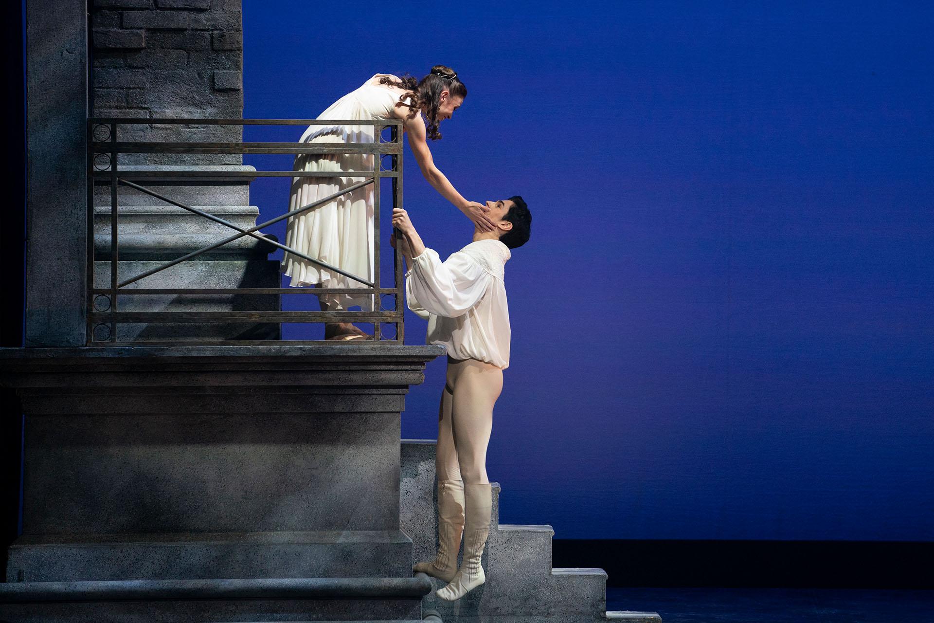 Wearing a white shift, a dancer leans over her balcony and lovingly hold the face of her love as he climbs up to see her