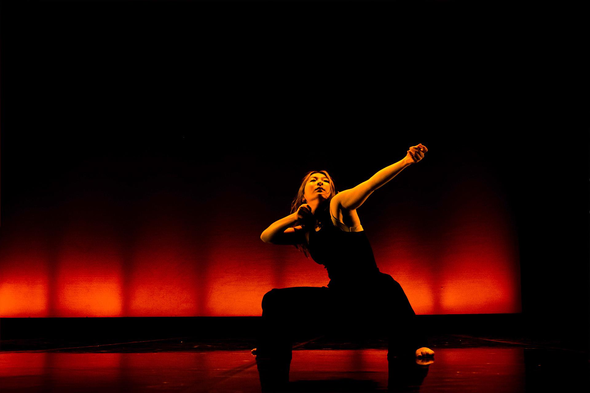 Dancer wearing black trousers and a black vest squat, one arm bent and the other extended as drawing an archery bow.