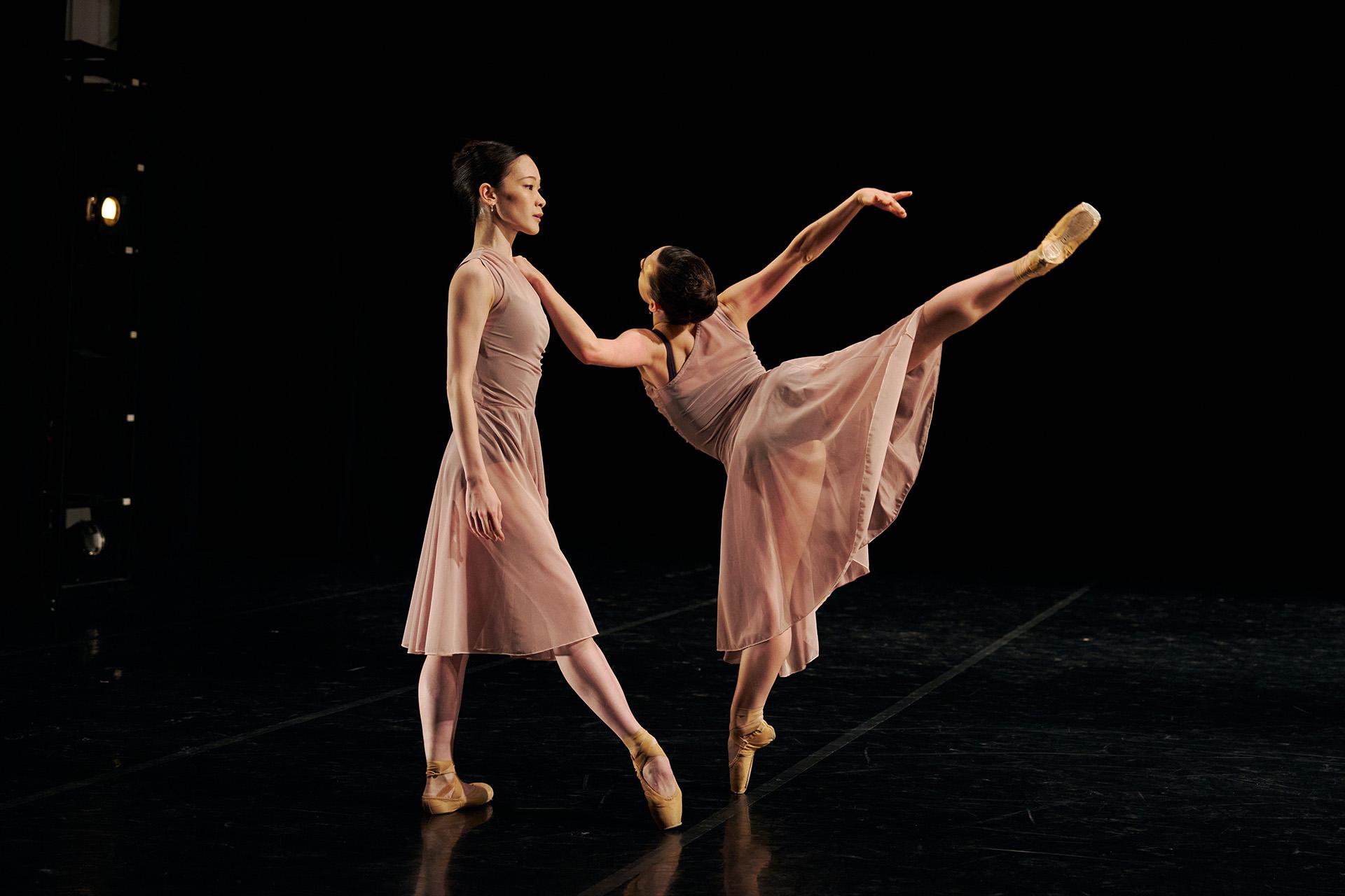 A dancer stands ambivalent while another holds her shoulder and poses en pointe arabesque