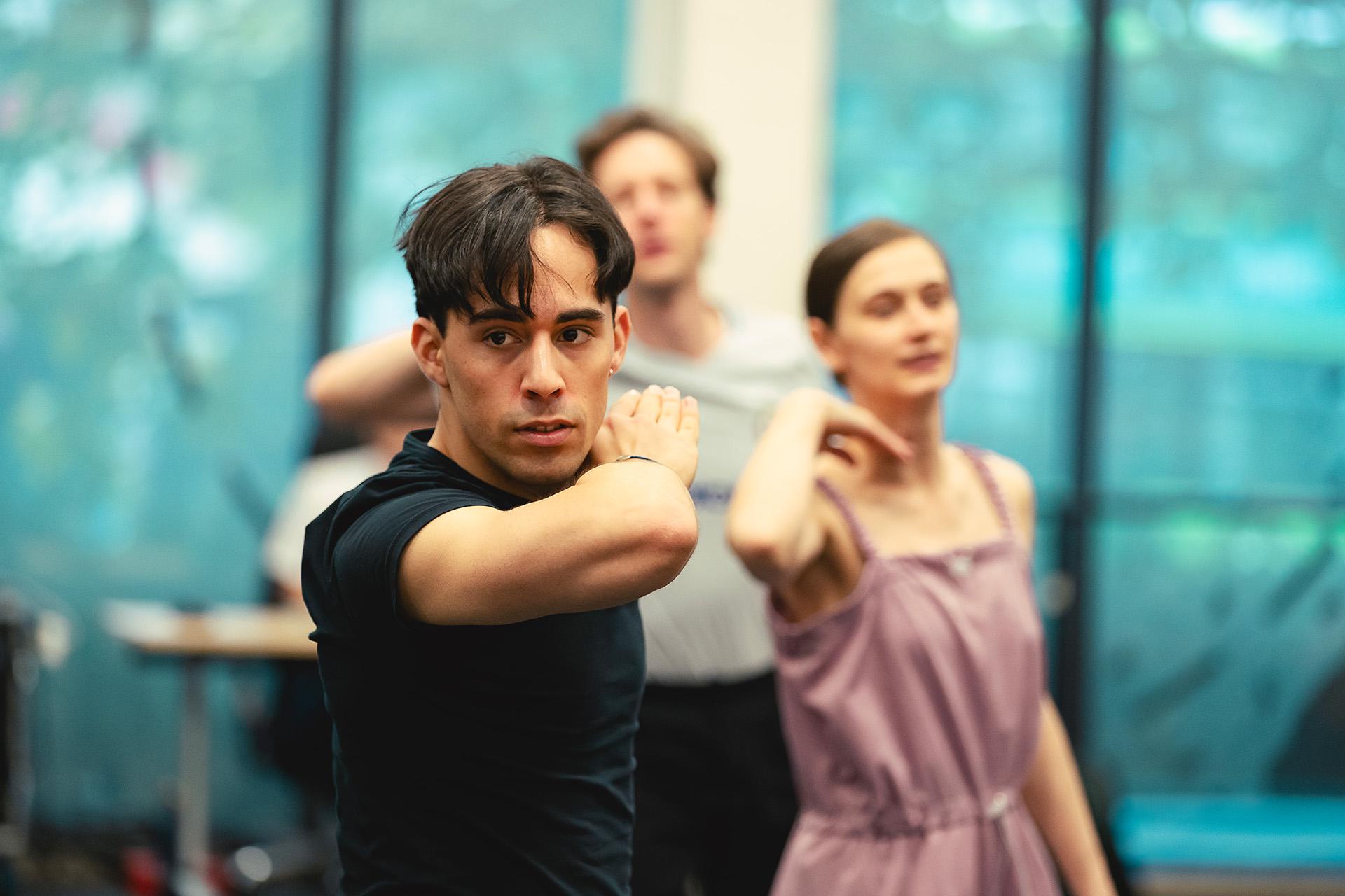 Dancer in a black T-shirt in rehearsals stands with his arm level with him face, folded in at the elbow