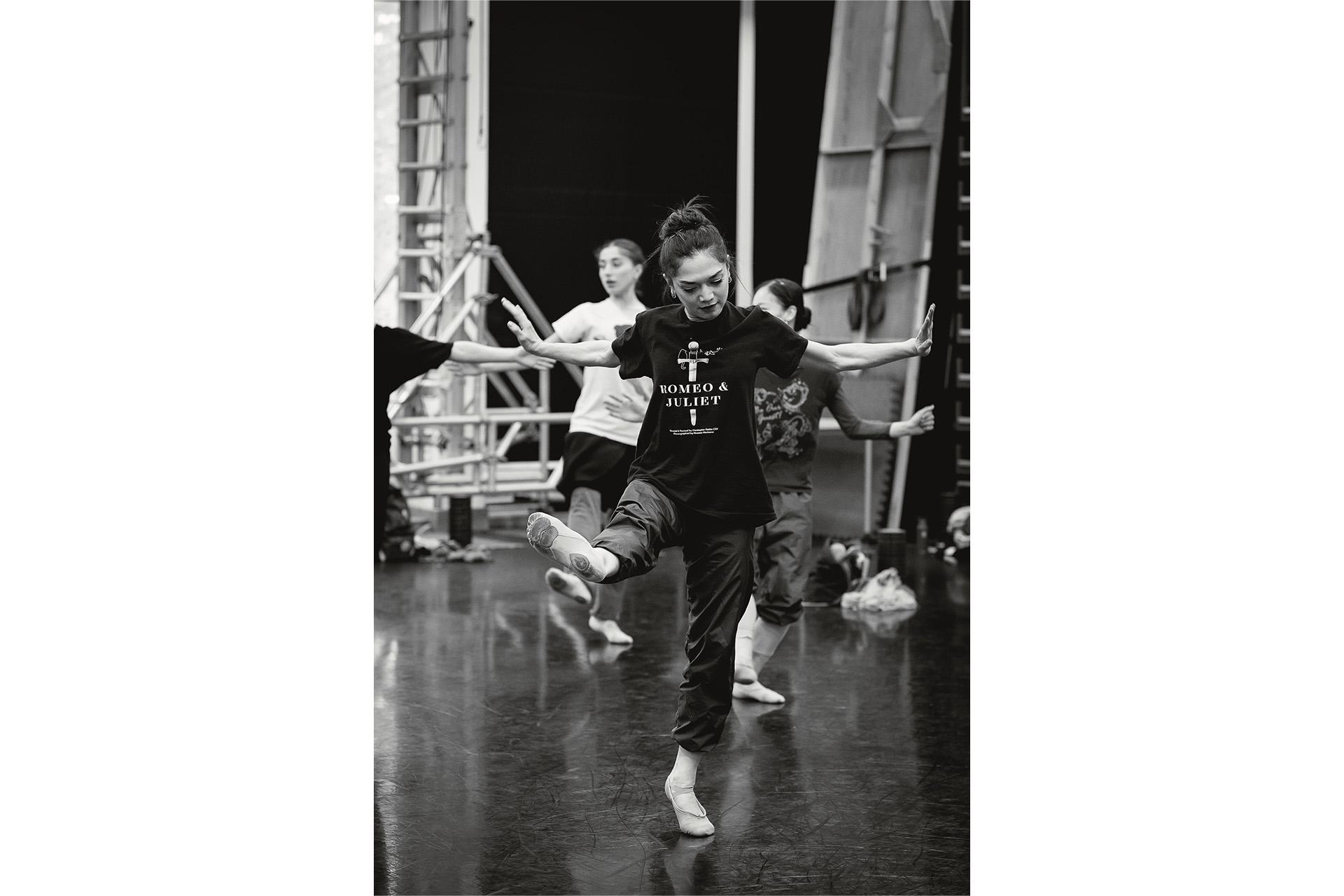 Black & white image, in rehearsal a dancer stands on one foot, the other extended to the front, her arms wide to the sides