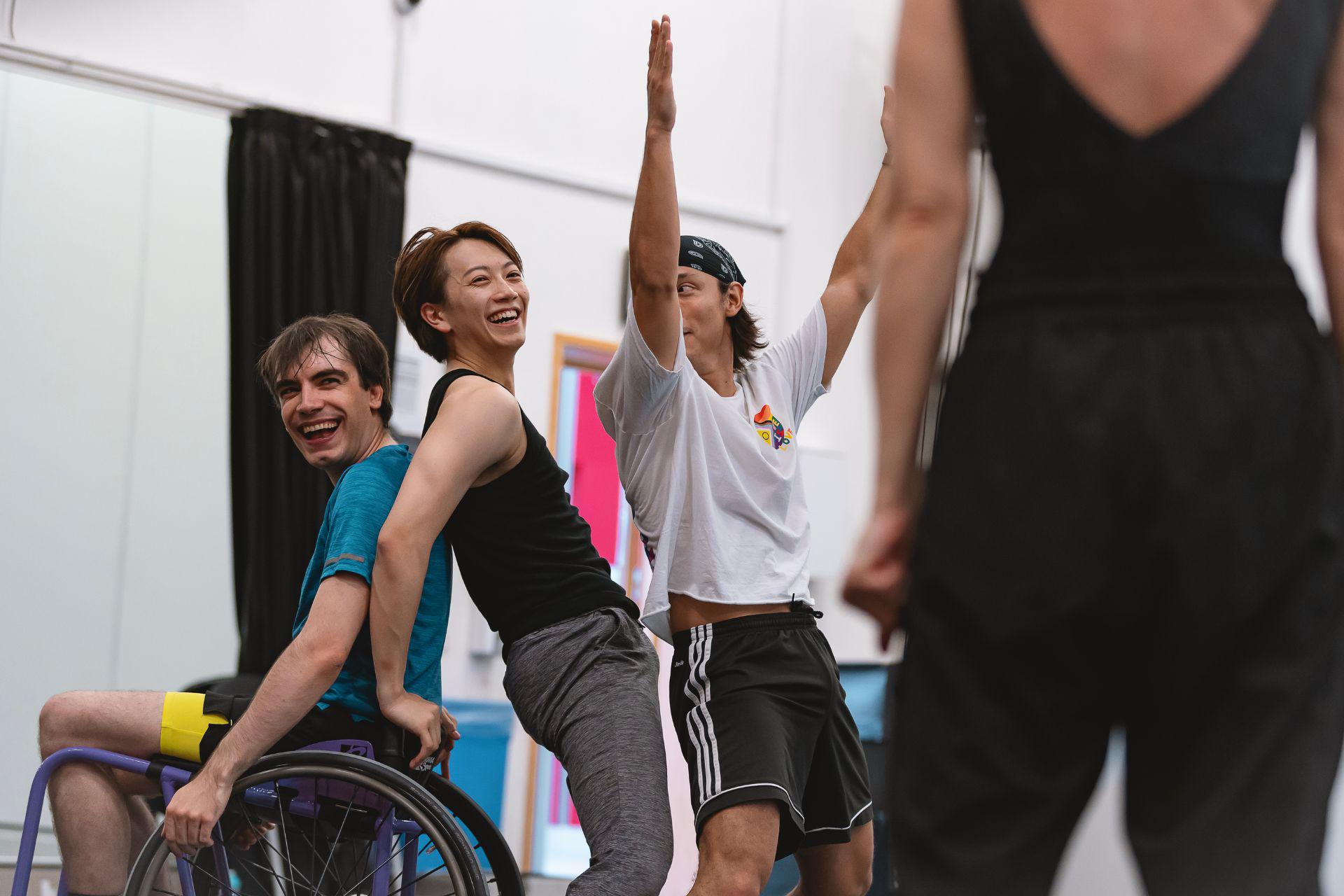 A wheelchair dancer laughs with another dancer who leans backwards, resting his hands on the wheelchair for support. Another dancer stands next to them with his arms in the air. The choreographer can be seen watching them in the foreground.