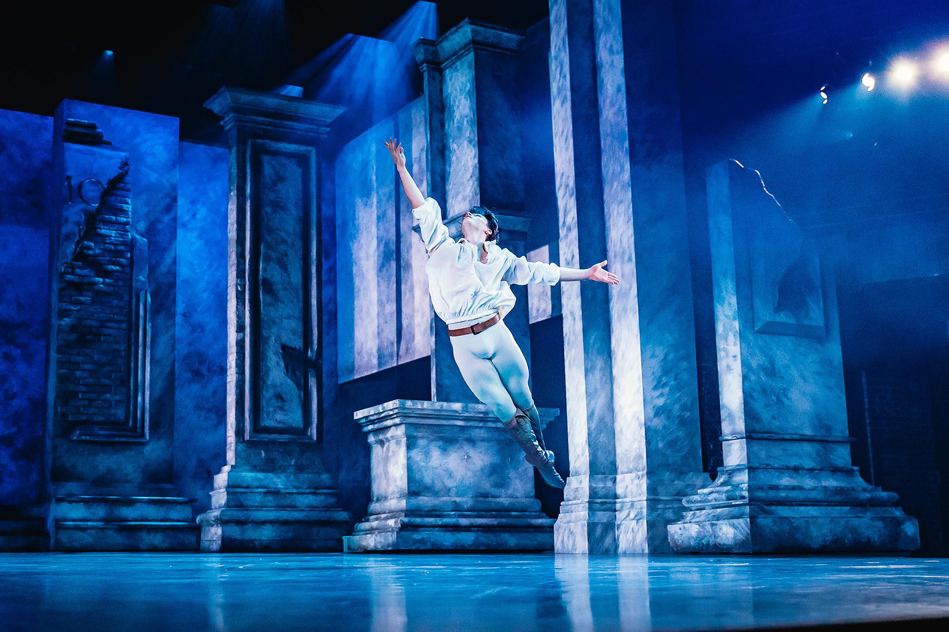 Romeo & Juliet, 2024, production, dancer Harris Beattie, photo Emily Nuttall Dancer leaping across the stage, his legs together, one arm raise triumphantly above him, bathed in blue light