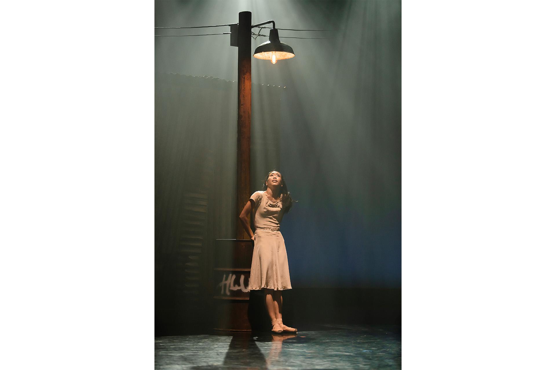 Dancer in light tan dress leaning against a lamppost illuminated by the old-fashioned light directly above her