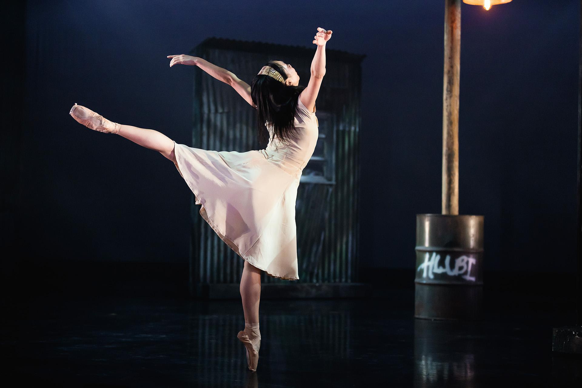 Dancer in a pale dress wearing a hair band stands on one foot en pointe, a leg straight behind her, both arms above her head