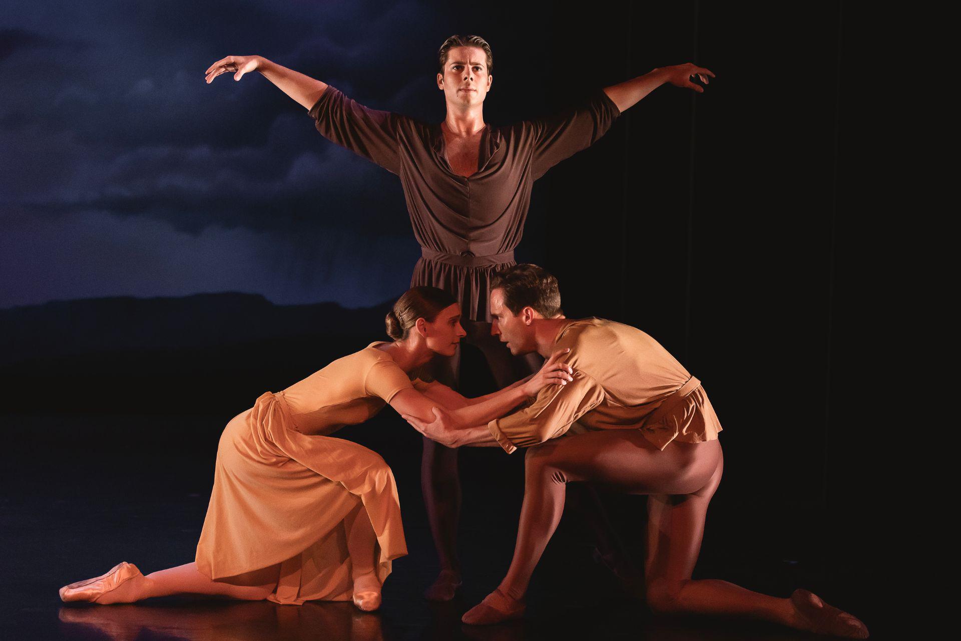 Two dancers kneel in front of each other, holding onto one another's arms and looking into each other's eyes. Another dancer in dark colour stands between them, their arms floating upwards.