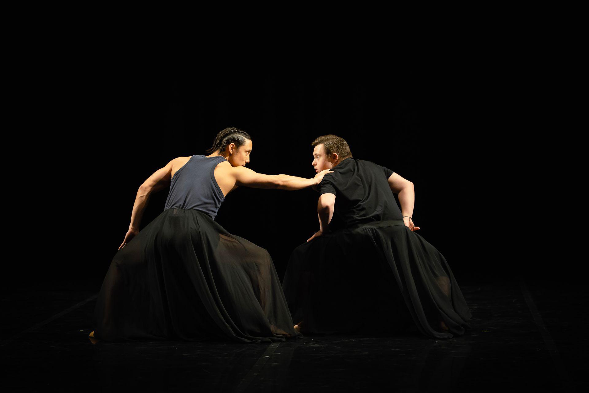 Two dancers stand hands on their bent knees. They look at each other as one grabs the other's shoulder.