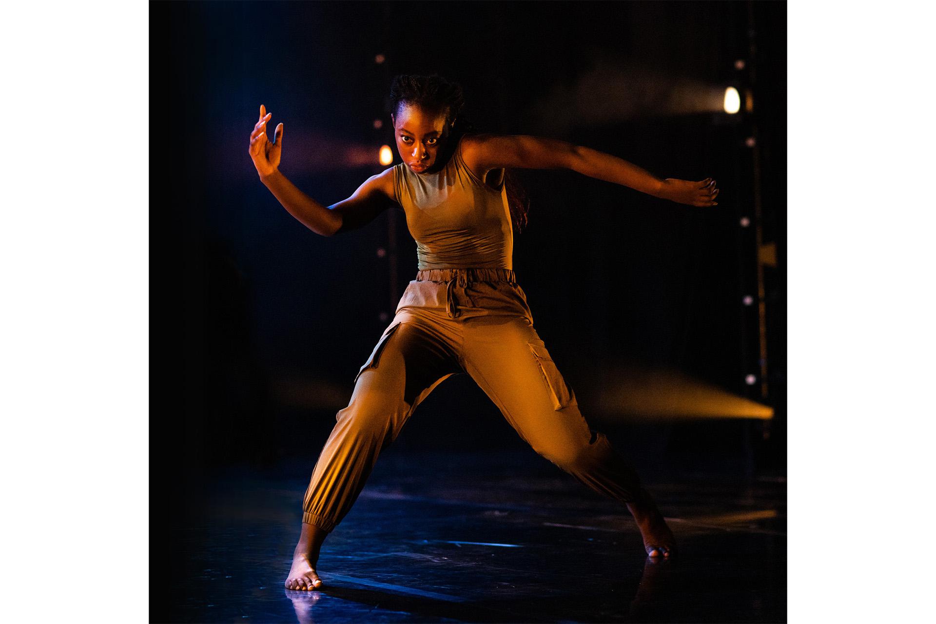 A dancer, legs apart and knees bent, her arms wide, looking straight at the camera