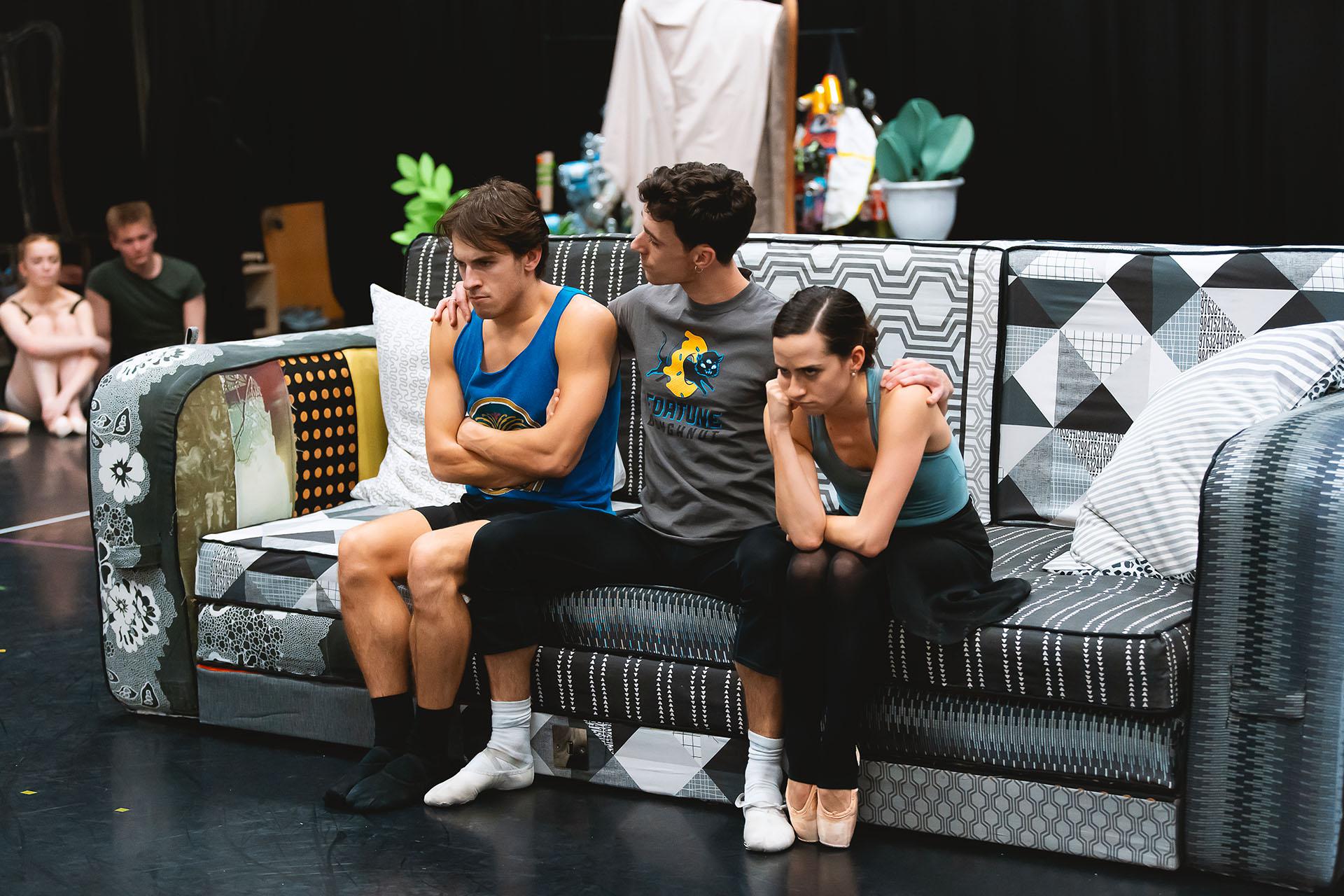Two dancers sit on a sofa looking very upset separated by a taller dancer looking caringly at them with a hand on their shoulder