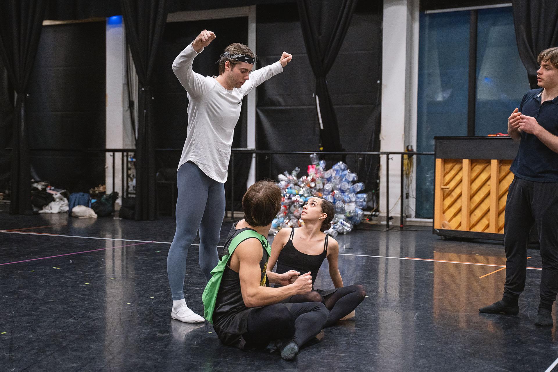 Two dancers sat cross-legged on the studio floor both looking behind them to see another looming over them