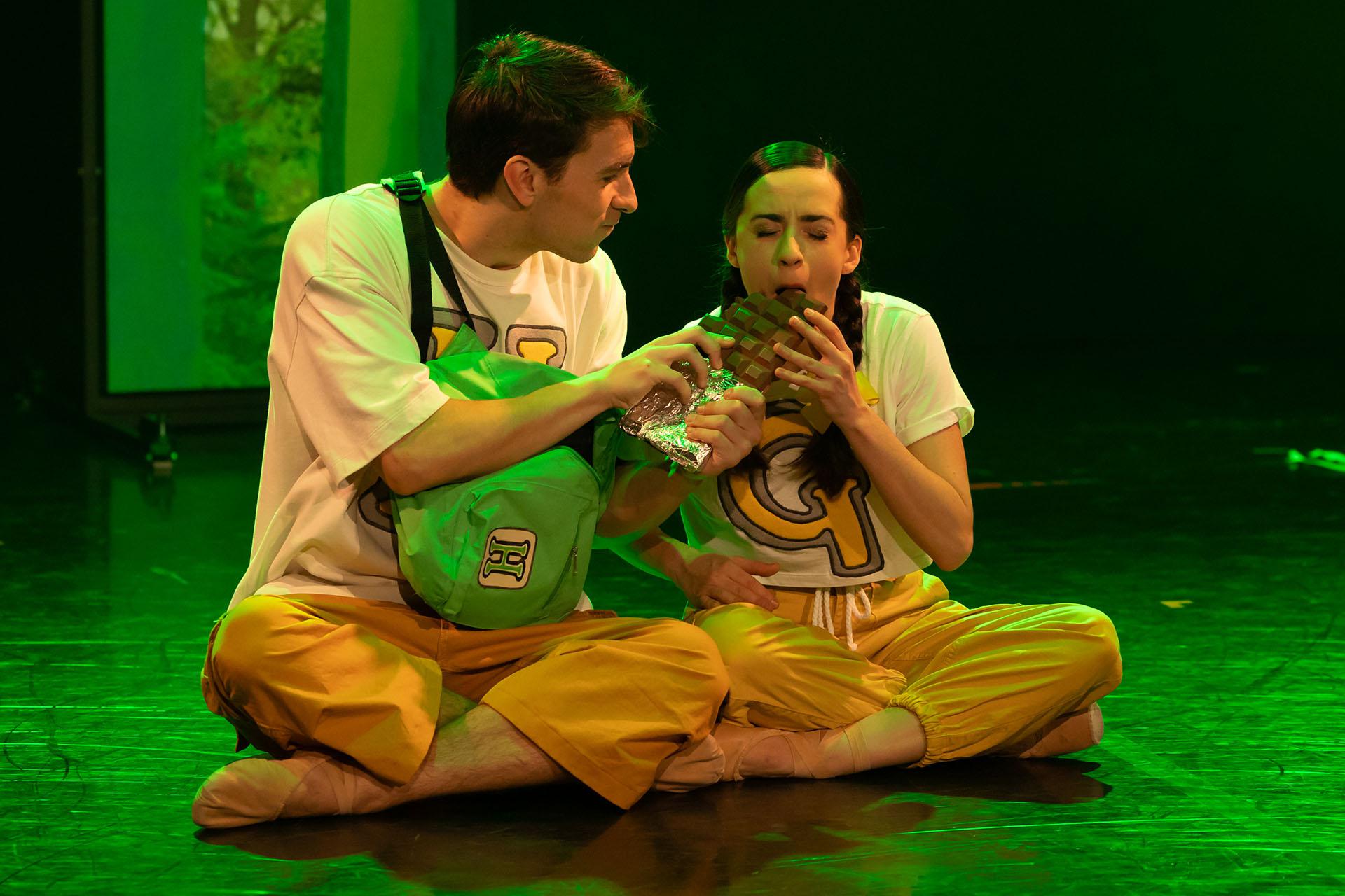 Hansel and Gretel sit on the floor in the forest, they're eating a big bar of chocolate.