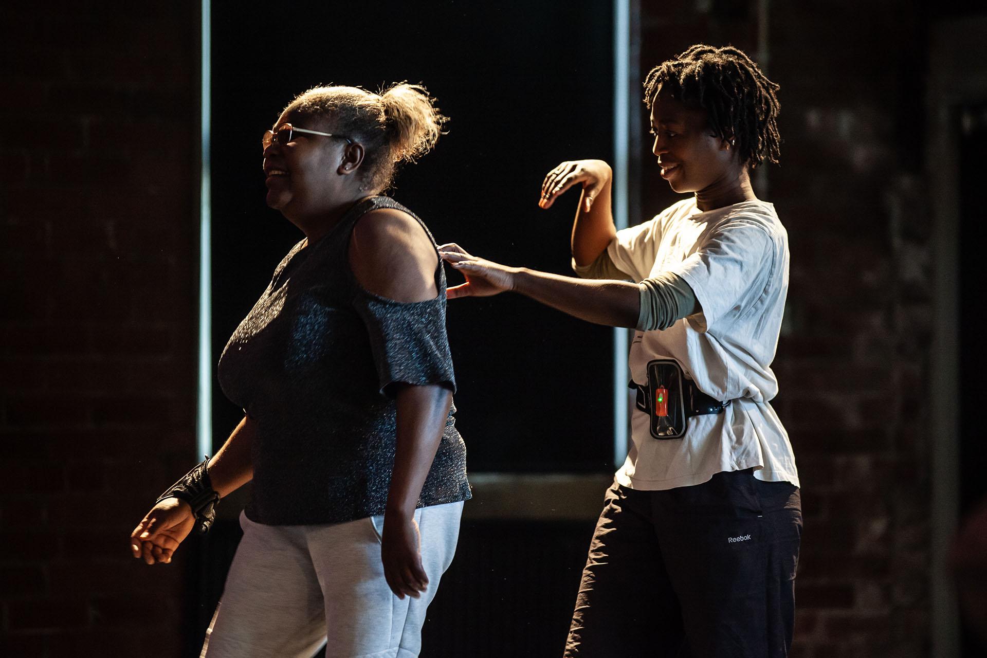 One person in front of the other, the person behind guiding the other by placing the fingers of their left hand on her back