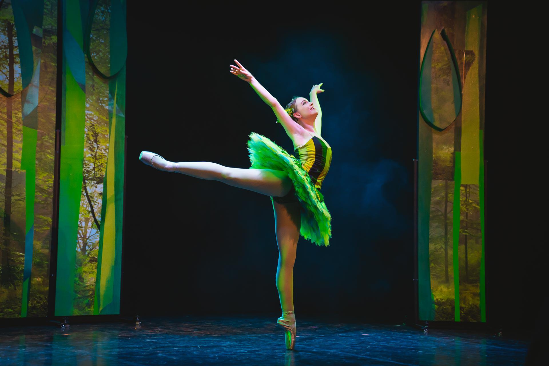 A dancer stood on pointe with arms extended and leg reaching back. She is wearing a vibrant green tutu and is standing in a forest background