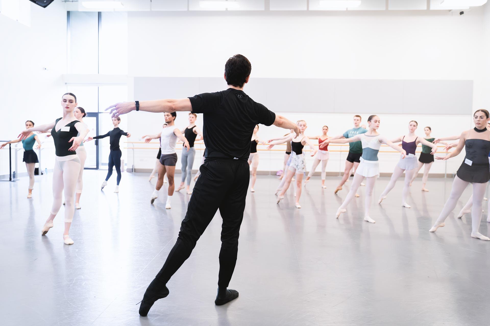 A teacher in all black stood facing away from the camera teaching a ballet class