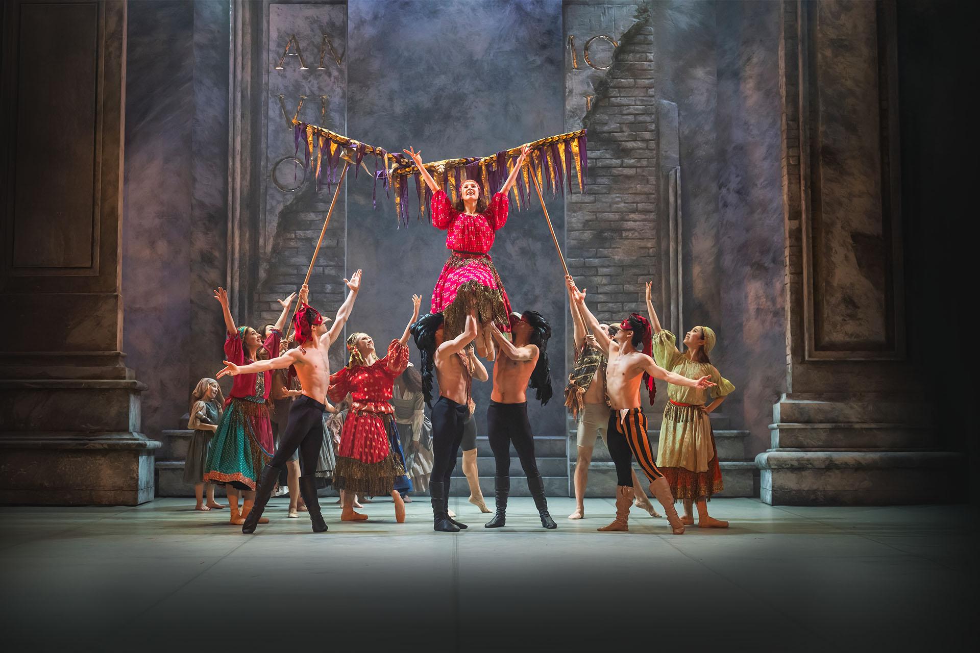 Men in masks and black leggings lift a woman in a pink patterned dress and she raises her arms in celebration