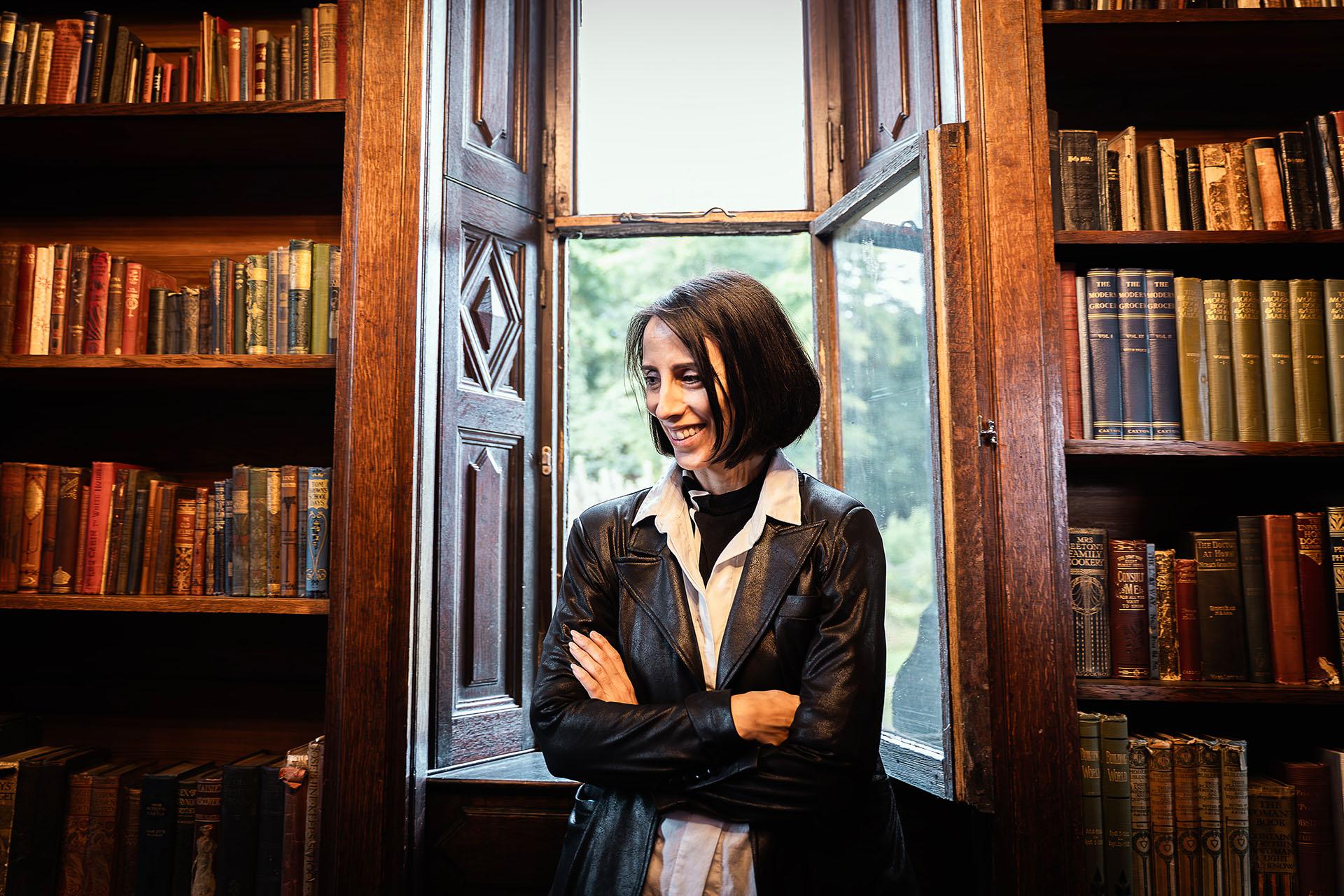 Annabelle Lopez Ochoa with dark brown hair, wearing a white shirt and black leather jacket, stood in front of a window in the Library at Shibden Hall and smiling.