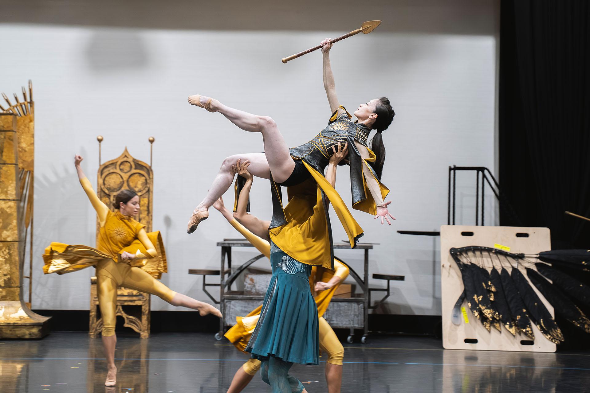 A woman warrior holds a lance above her while being lifted aloft