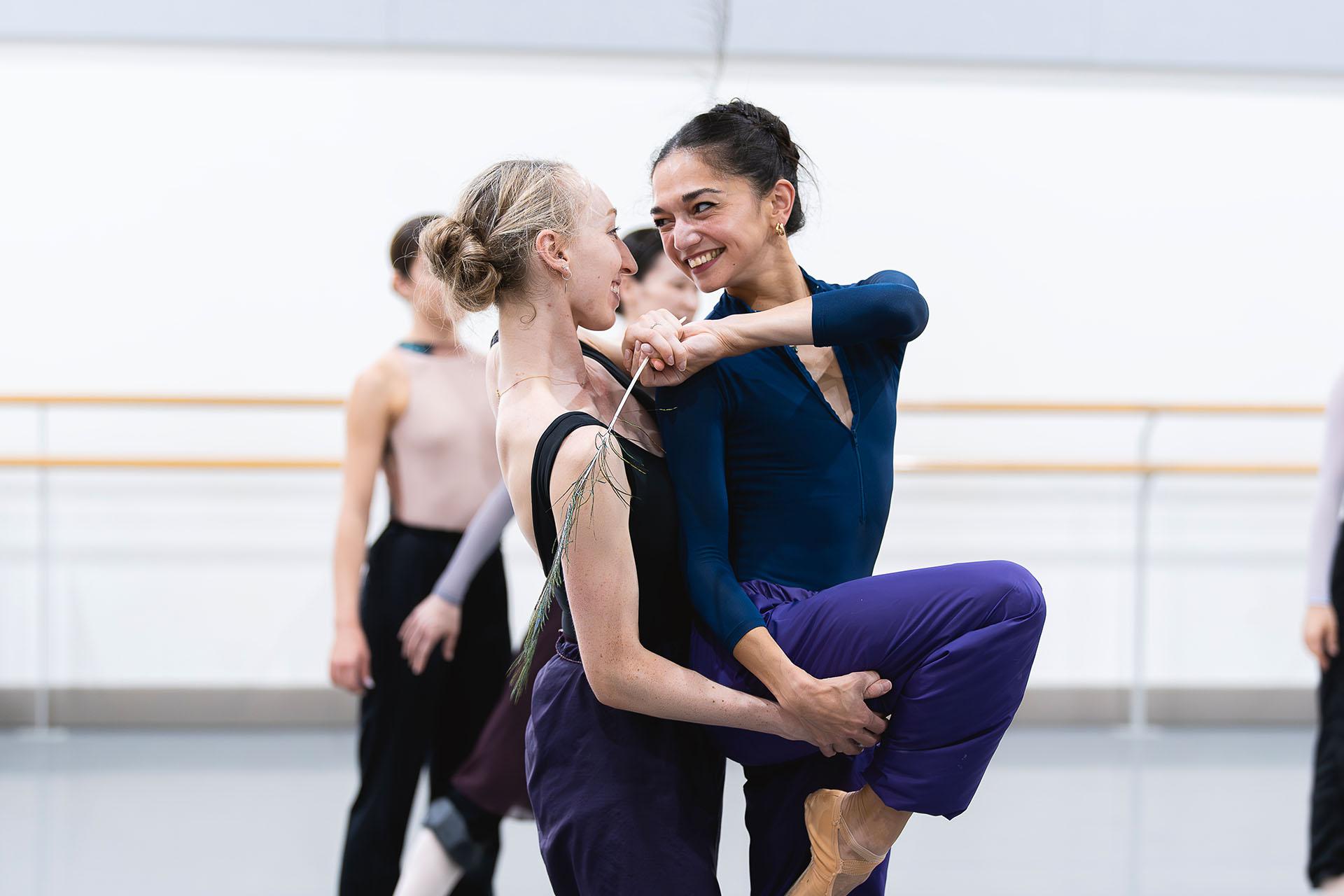 Two dancers looking at one another smiling, the one behind holding the other's leg at the thigh, the one on front reaching around playfully