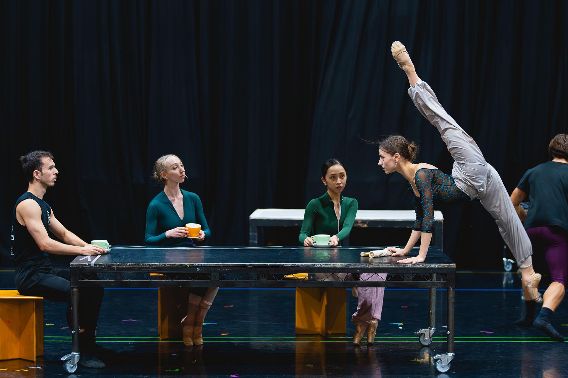 Four dancers around a table, three of them sat looking at the fourth whose palms are on the table, feet off the ground, one leg pointing down, the other opposite