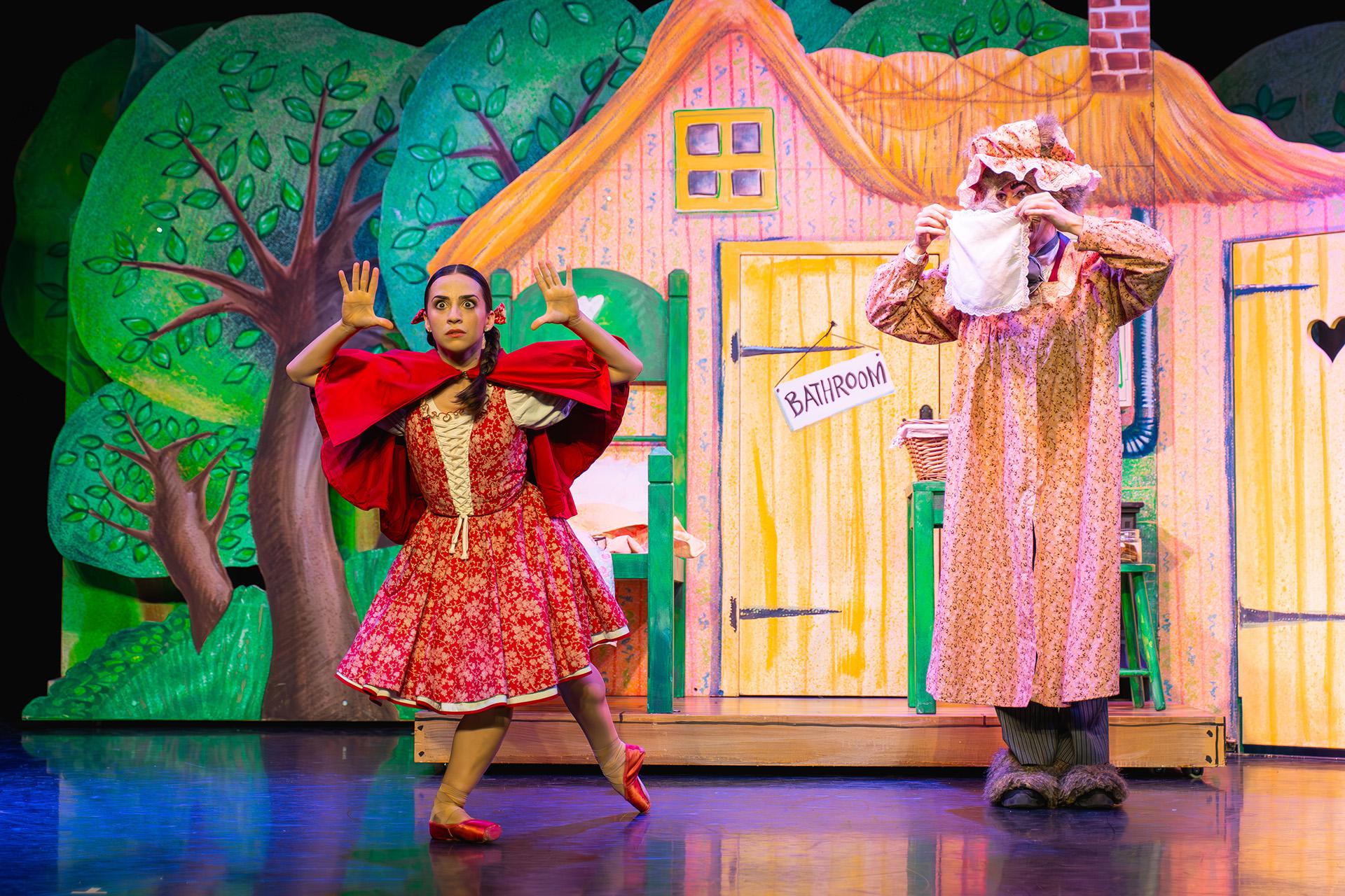 Little Red looks out worriedly while behind her the wolf dressed as Little Red's grand-mother hides their face behind a handkerchief