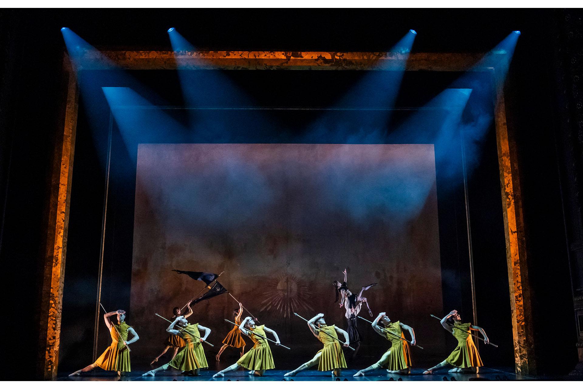 Chafts of light shine down on the army wearing yellow and carrying pikes, all standing in the same pose shielding their eyes