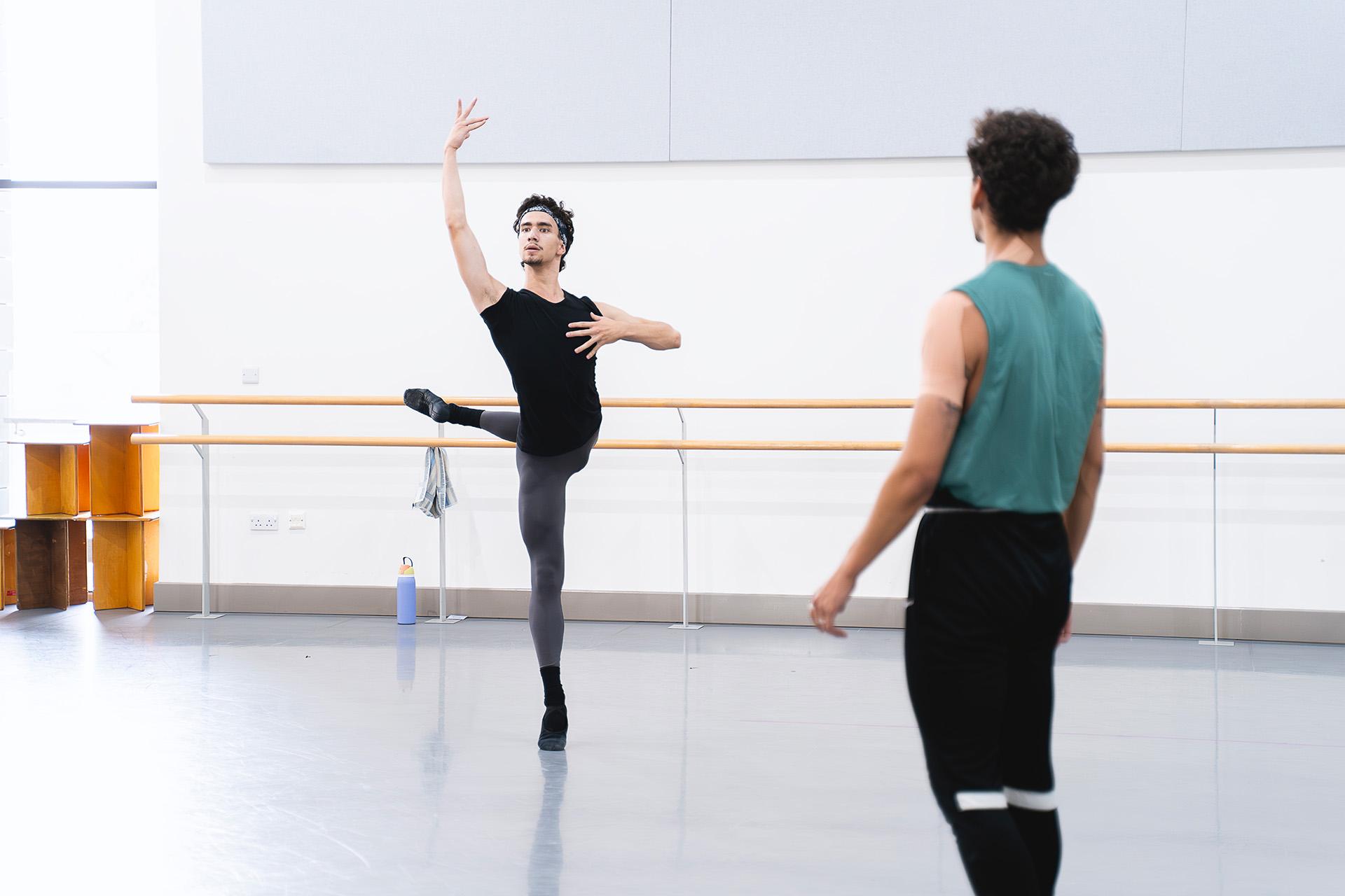 Young male dancer stands on one foot, his other leg raised behind him while someone looks on