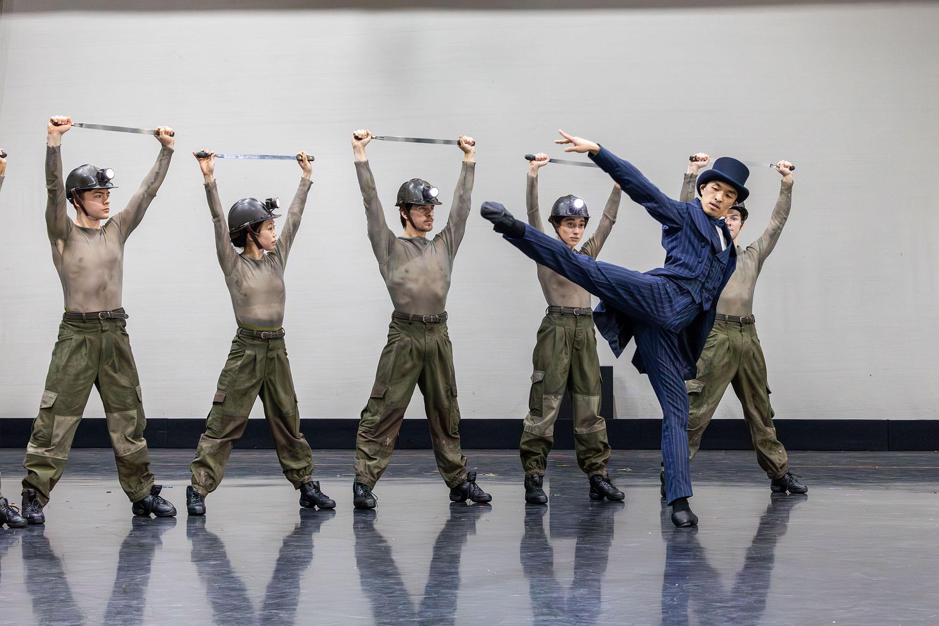 A man in a dark blue suit dances in front of workers in drab uniforms and miners helmets
