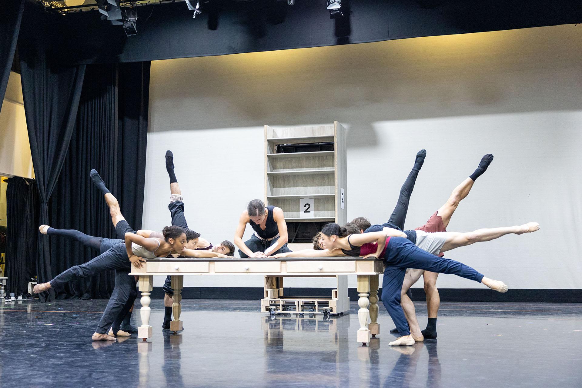 A dancer in black sits on a wooden table, leaning over whilst writing with a feather quill. Multiple dancers surround the table, each with one leg elevated behind them, reaching towards the quill.