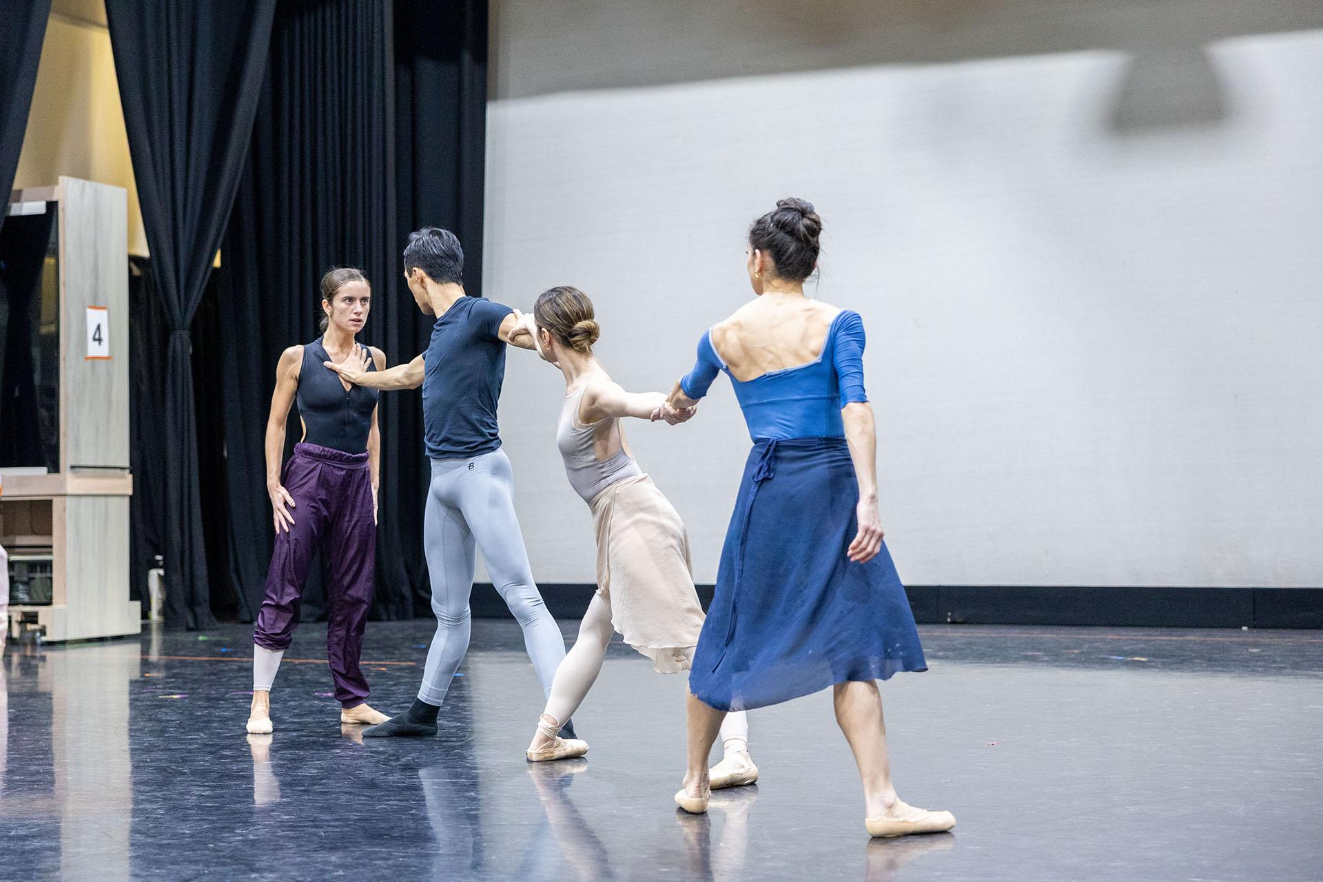 Three dancers stand in a line, holding hands; the one at the end reaches forwards, pressing a palm against another dancer's chest.