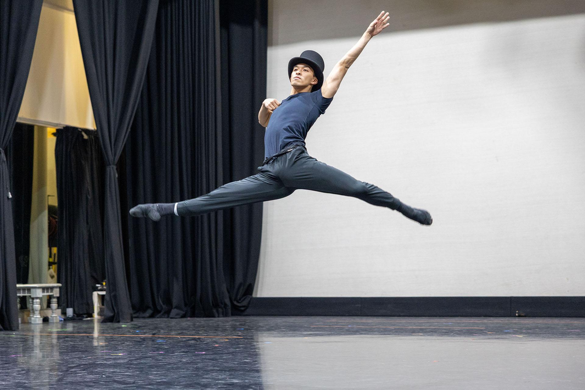 A male dancer in a top hat leaps a grand jete, one hand on his chest nobly and the other raised above his head