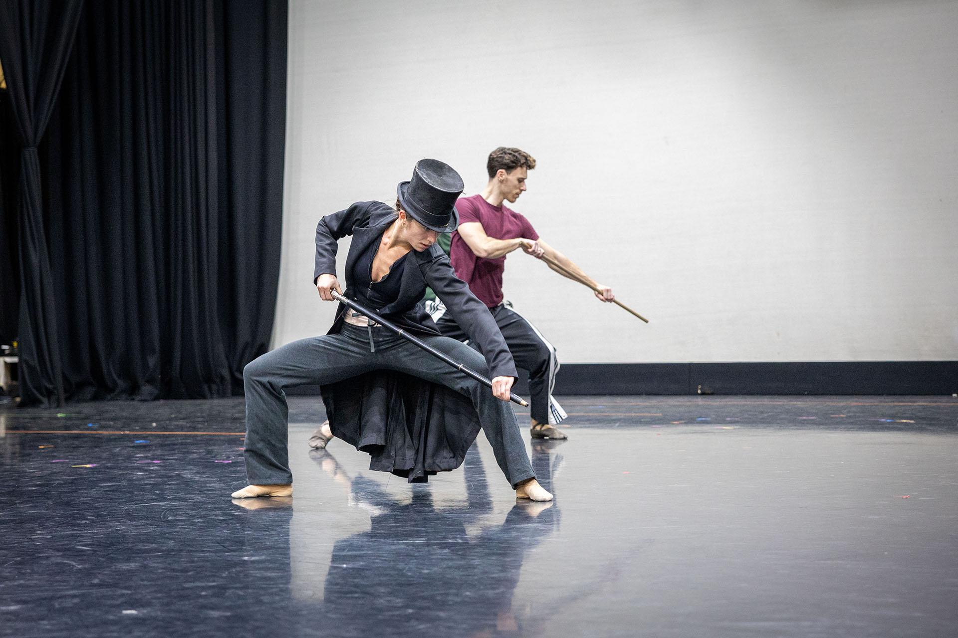 Rehearsing the role of Anne Lister, a female dancer wearing a long black coat and top hat stands, knees bent almost in a crouch, she looks down hiding her full face