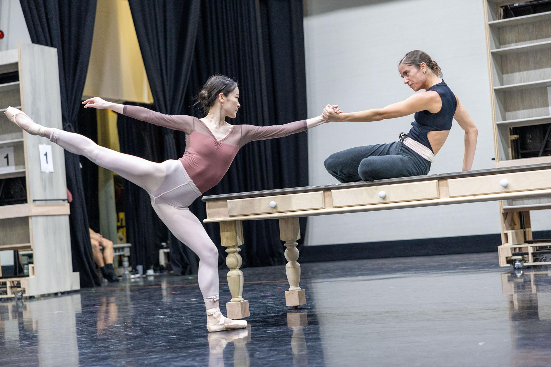 A dancer in black sits on a large wooden table. She holds hands with another dancer in pink, who has one leg elevated behind her. They both lean back, balancing their weight with each other.