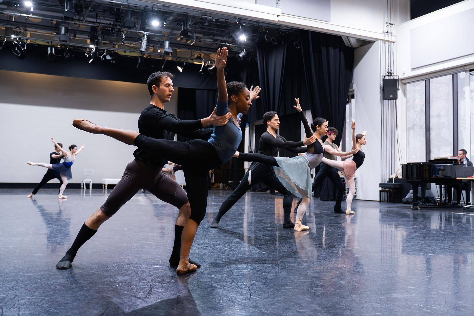 Three pair of dancers, the woman on one foot, a leg behind her, arms raised, a male dancer holding her stead just under her arms