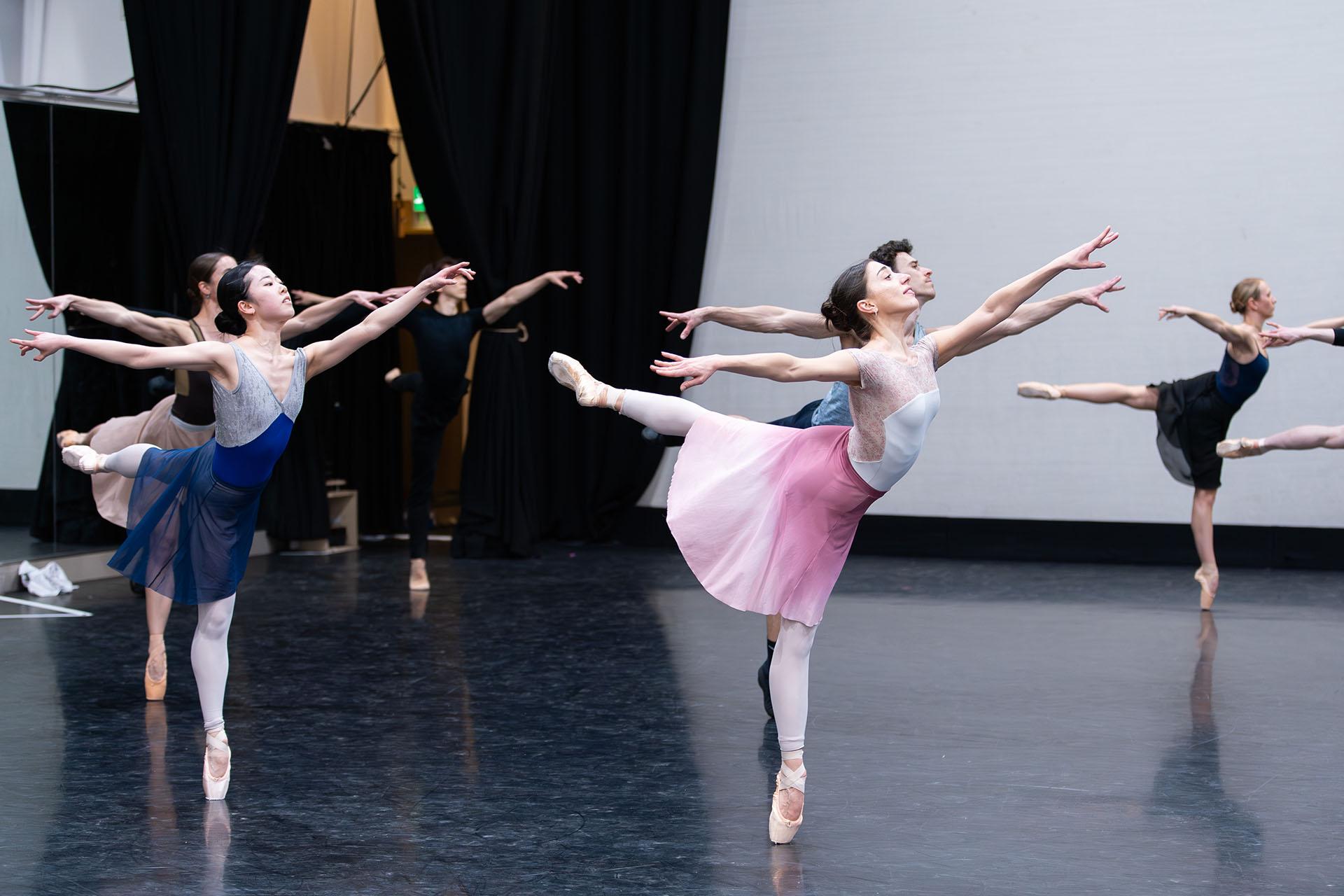 dancers each arabesque, in the foreground a woman in white with a pink hued skirt