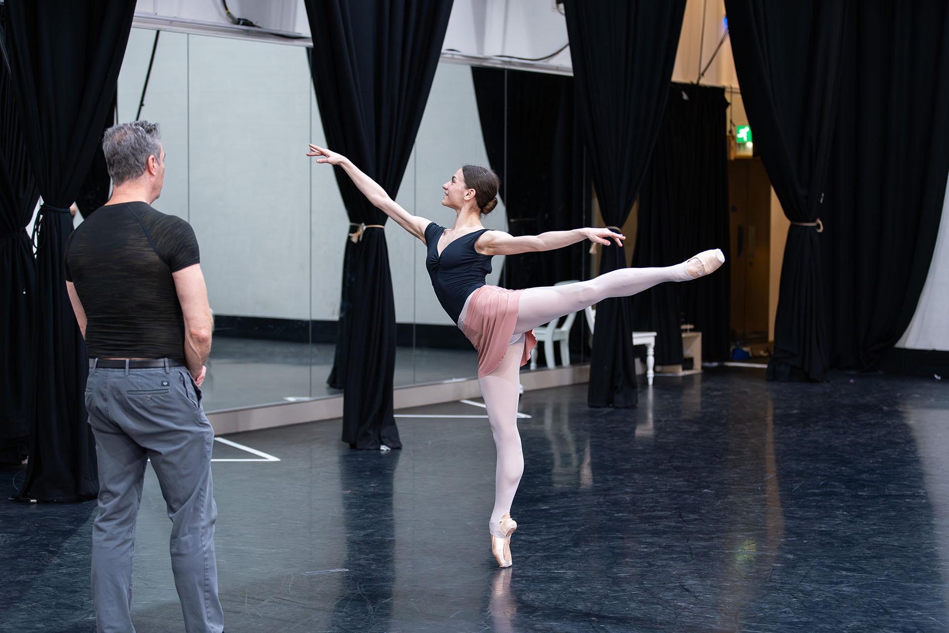 The choreographer looks on, coaching the dancer in the rehearsal room as she stands arabesque - one leg stright behind her, the other en pointe on the dance floor