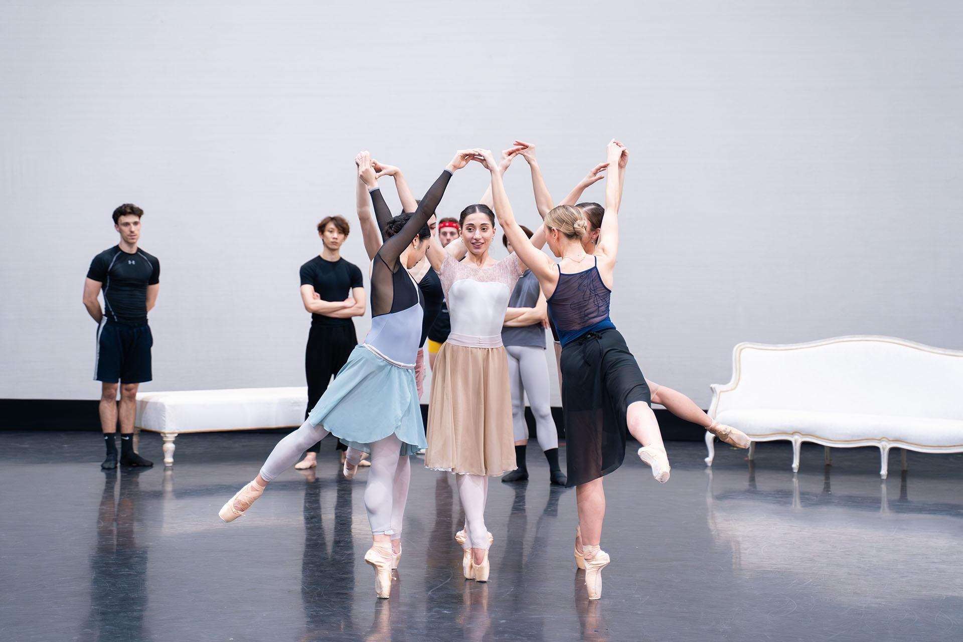 In the rehearsal room, dancers in a circle, their arms raised, hands touching each others, all on one foot en pointe