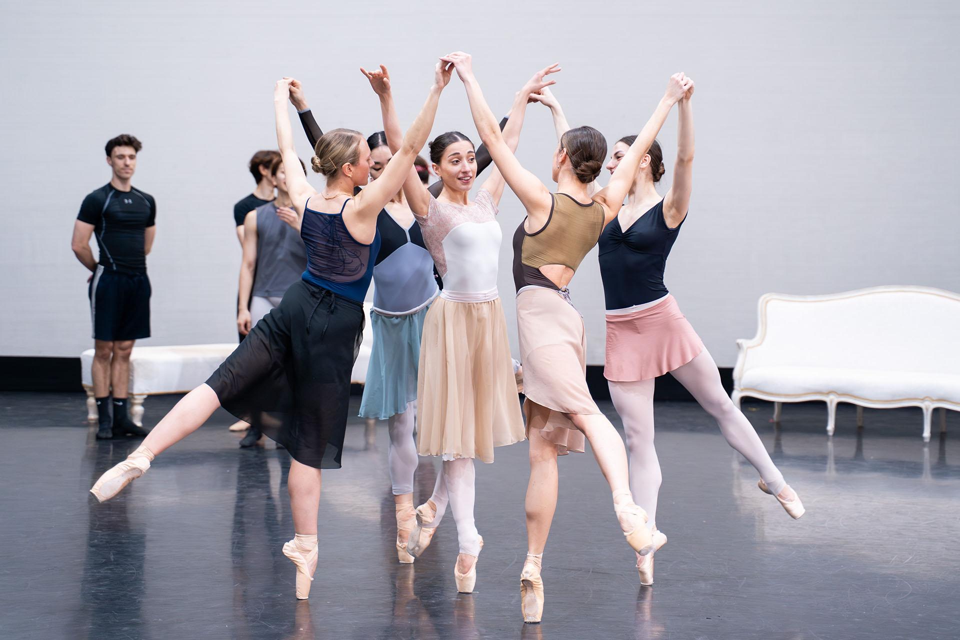 One dancer standing en pointe in a circle of fellow dancers, all on one foot, arms above their heads