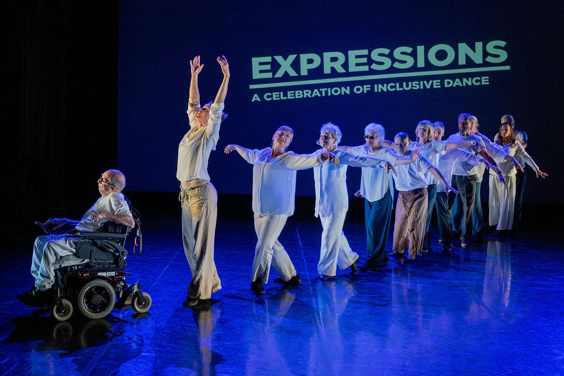 Dancers in white forming a row, each behind the other, arms wide; the person in from in a powered wheelchair