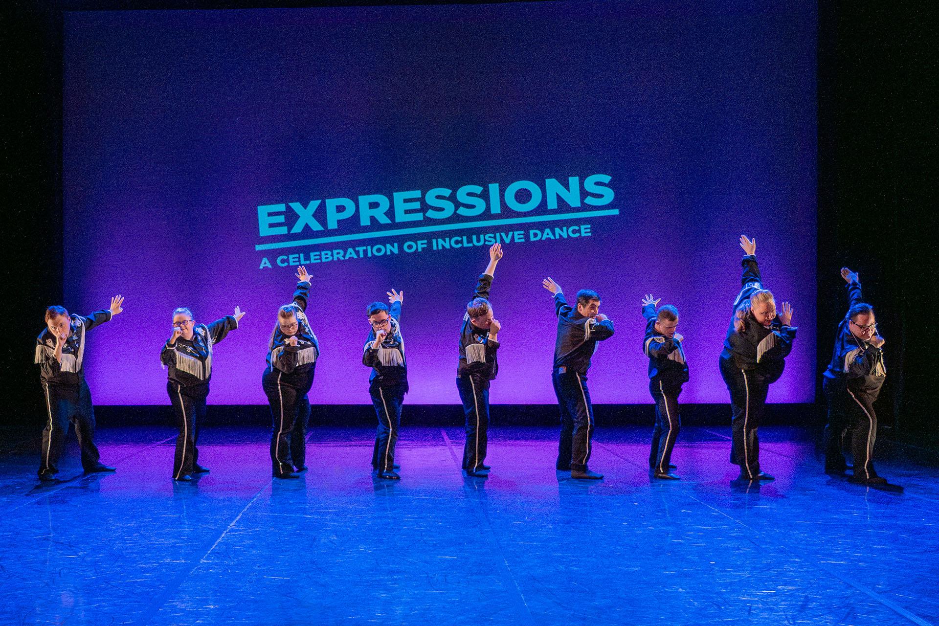 Nine dancers in tasselled shirts, one arm raised behind them, the other held to their mouth as if they're holding a microphone, posing in the style of a Las Vegas crooner