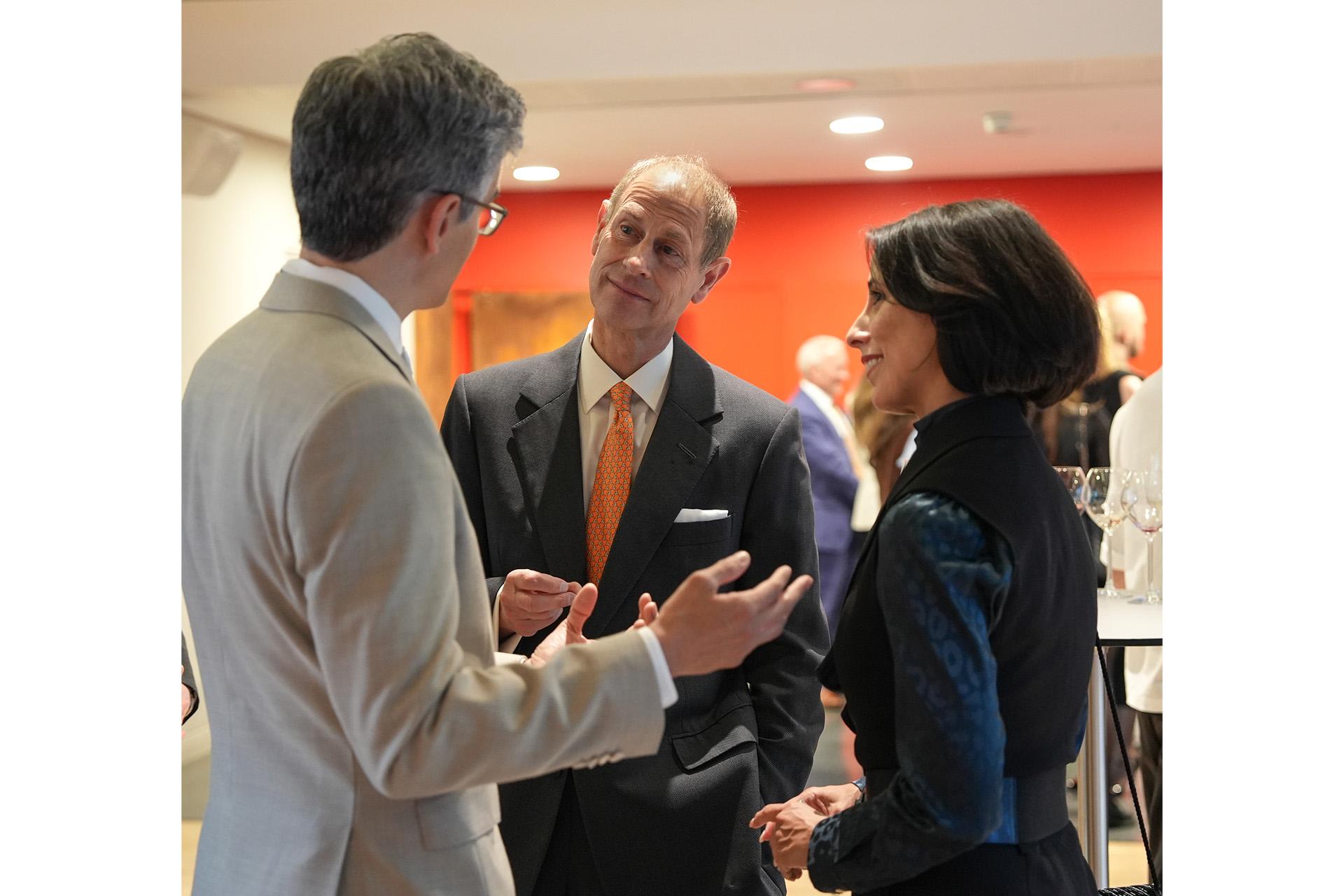 Man in dark quit and orange tie meeting another in a light suit and a woman in dark clothes.