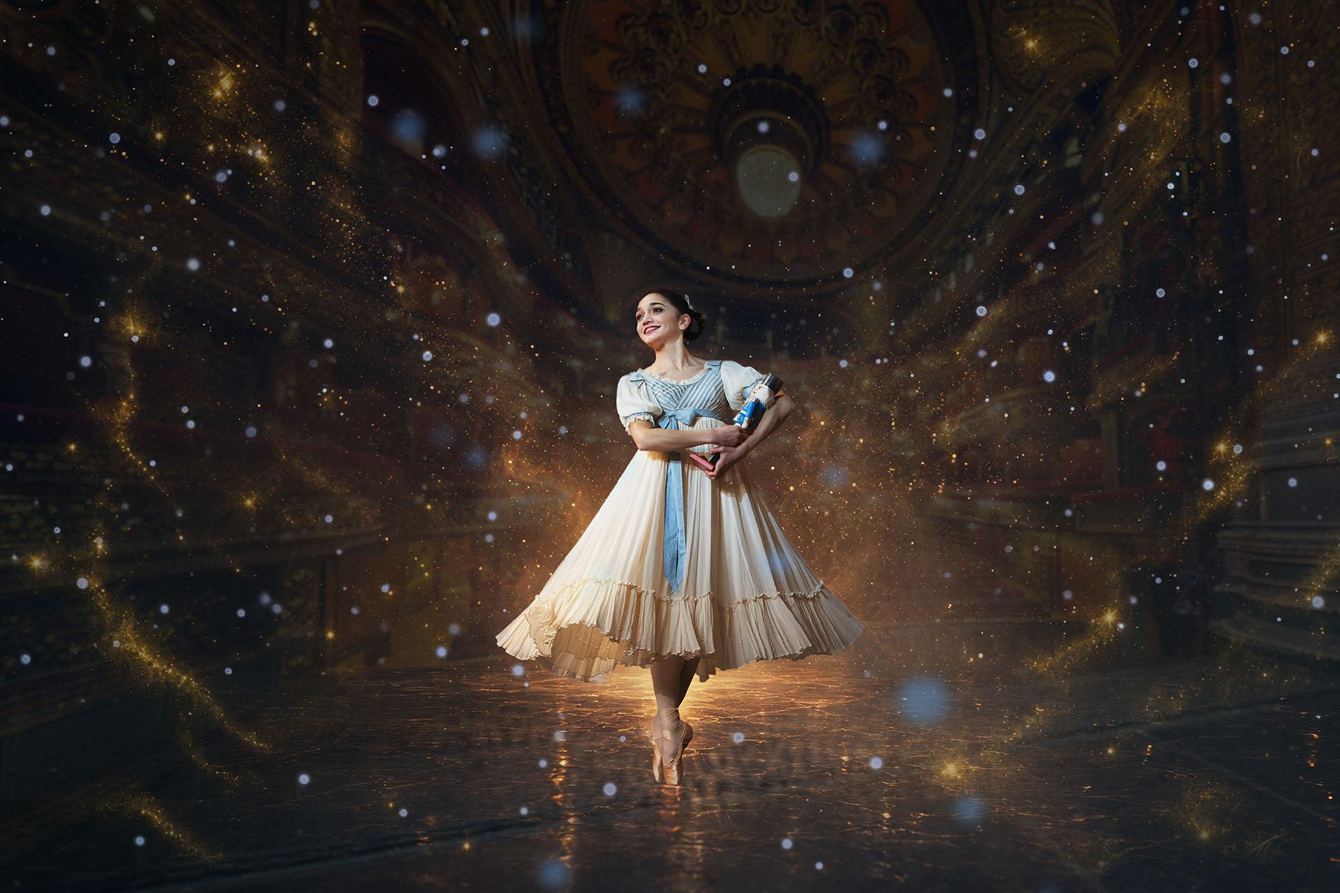 A dancer in Edwardian costume hold a Nutcracker doll in her arms, stands en pointe and smiles with childlike wonder