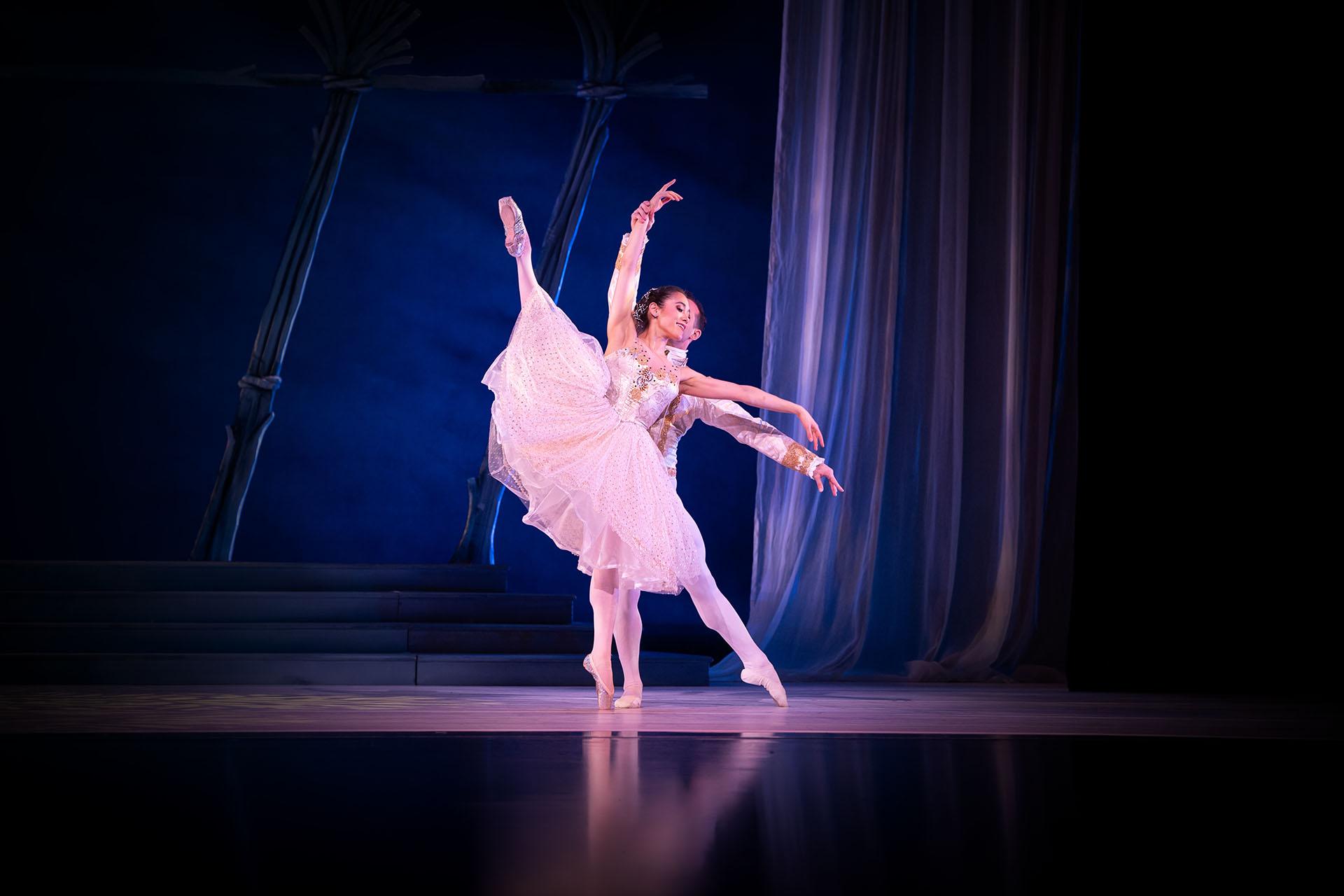 Woman in a shite flowing skirt and satin top stands on one foot en pointe and the other leg is exteded into the air, another dancer stands behind her supporting her balance.